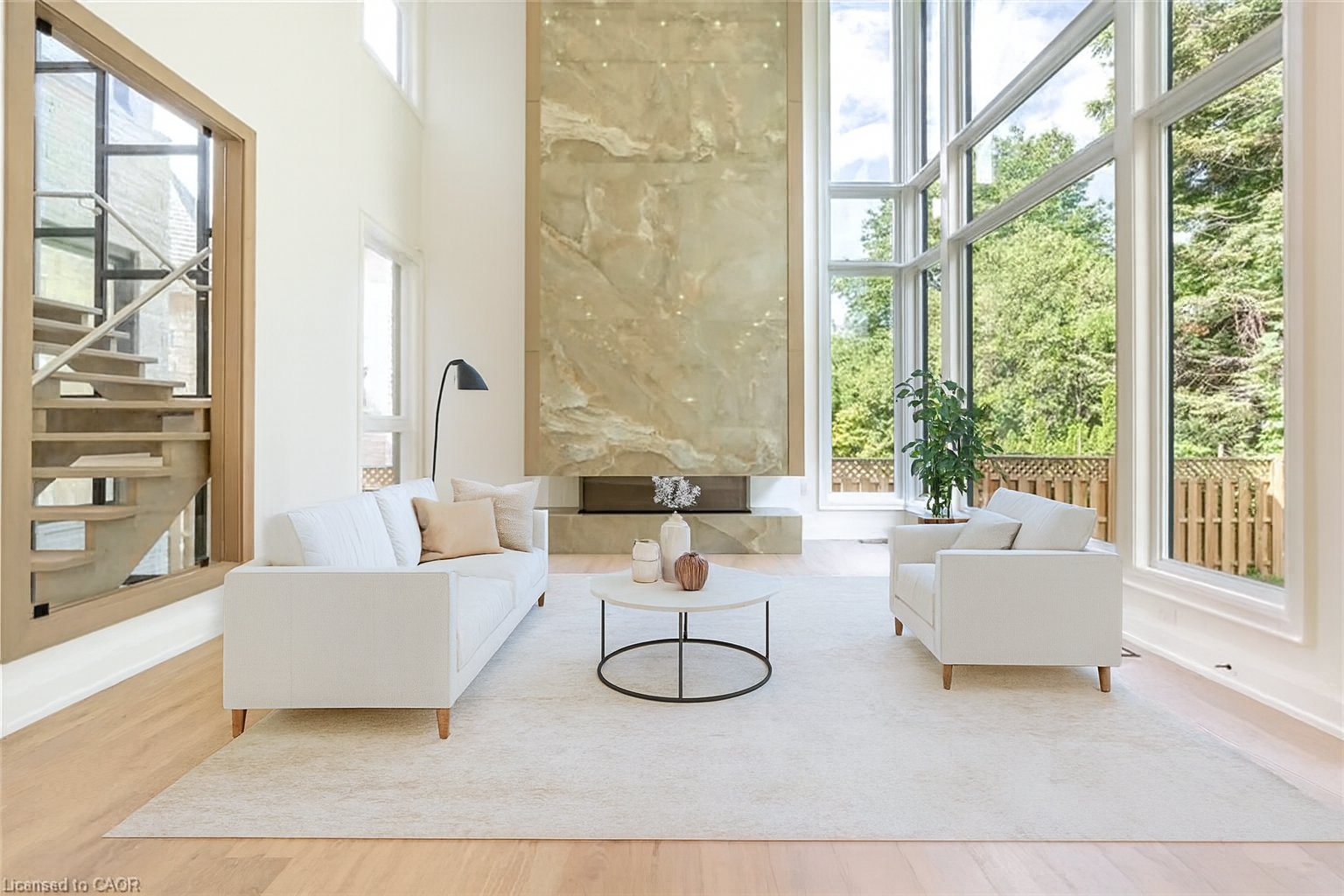 Living room with white sofas, a round coffee table with vases, large windows showing greenery outside, a tall marble fireplace, a black wall lamp, and a wooden staircase.