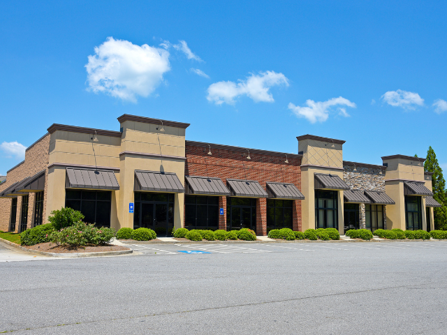 Empty shopping center building with storefronts, small bushes, and a parking lot under a bright blue sky. Commercial Property with shops