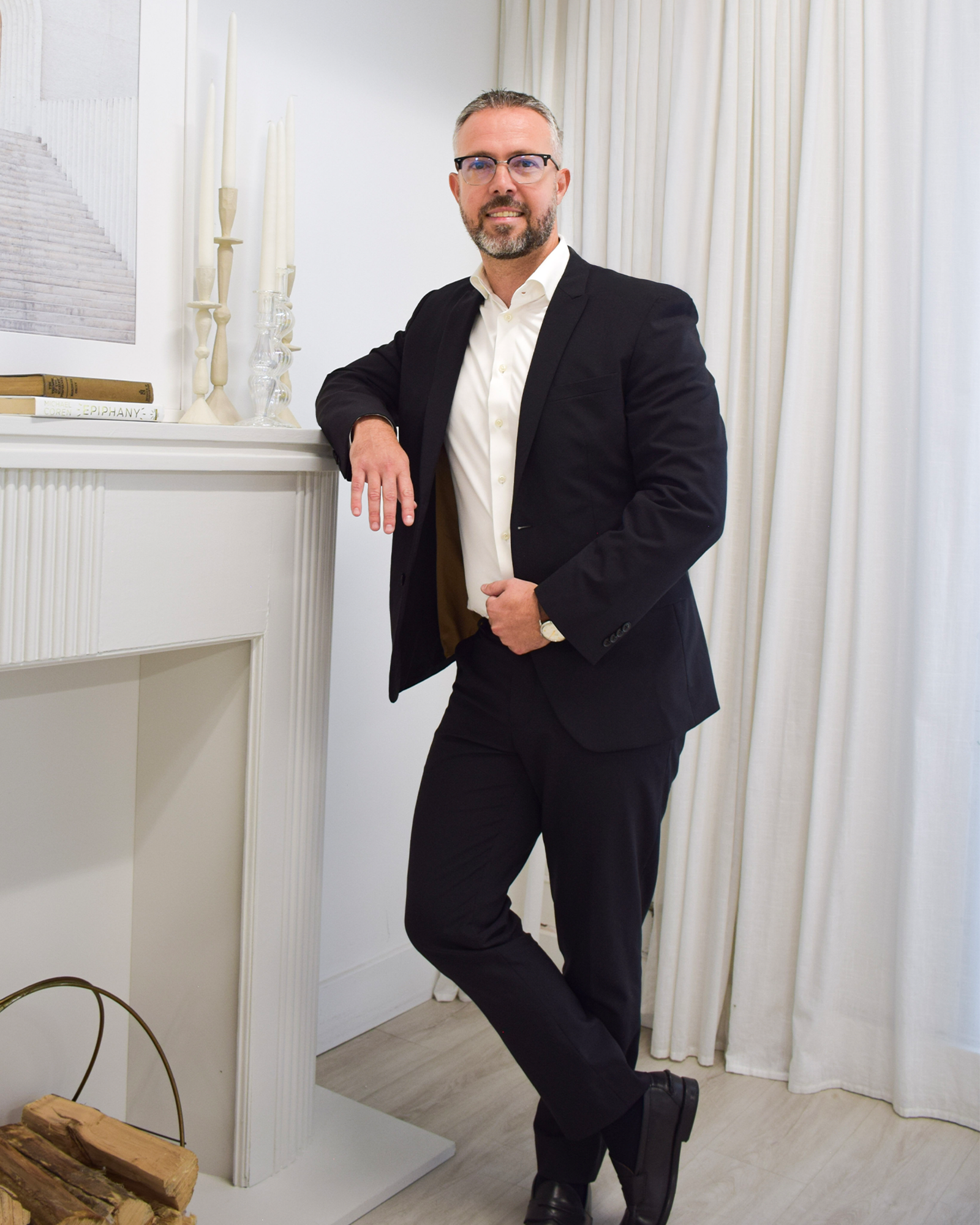 A man in a black suit and white shirt standing next to a white fireplace, leaning with one arm on it, in a room with white drapes and decor.