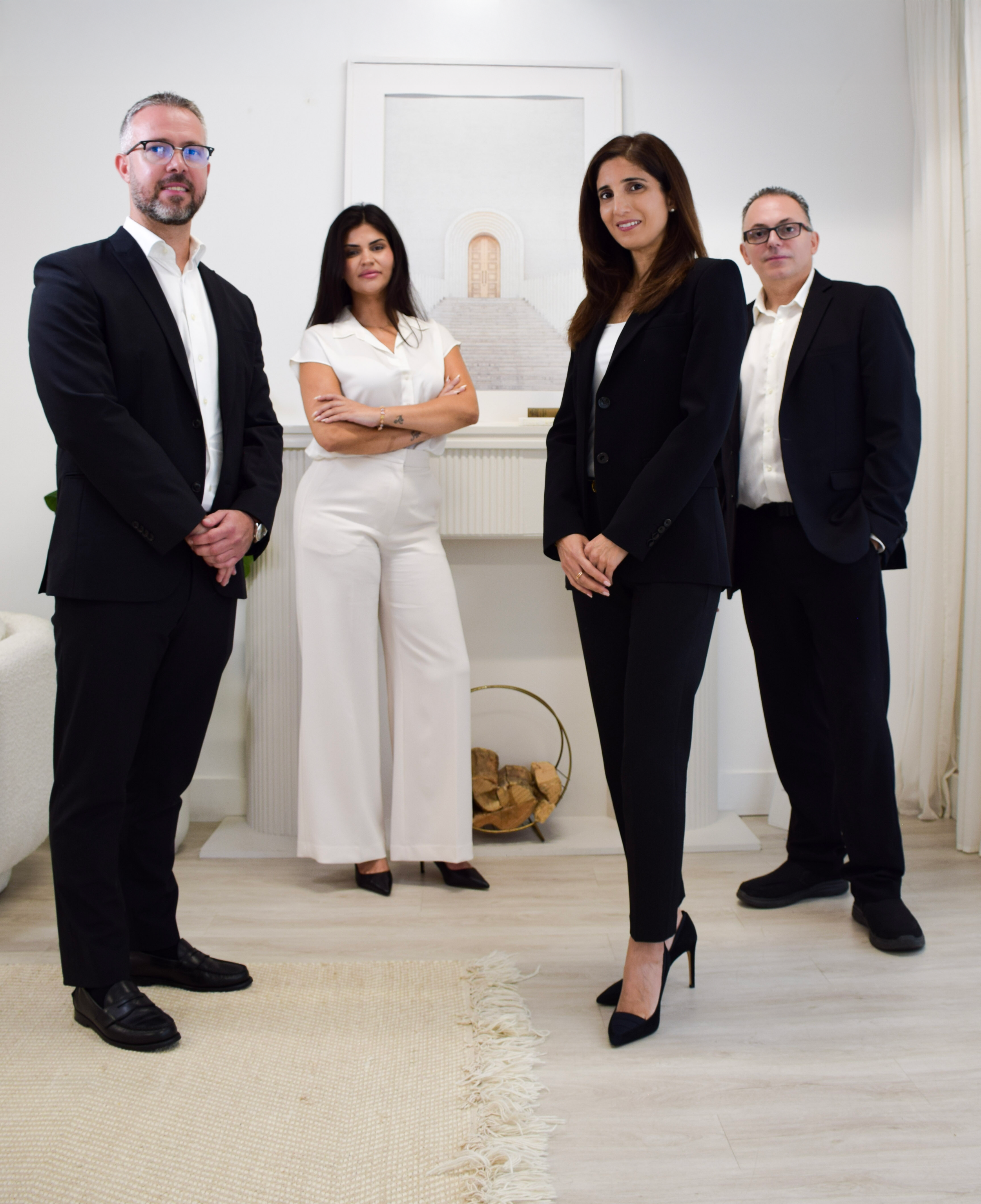 Group of four professionals in formal attire posing in a bright, modern room with art and a fireplace.