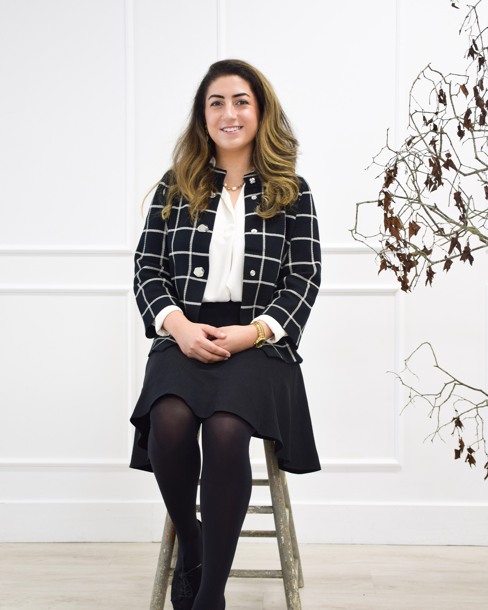 A woman sitting on a wooden stool indoors, wearing a black and white checkered blazer, white blouse, black skirt, and black tights, with a decorative plant in the background.