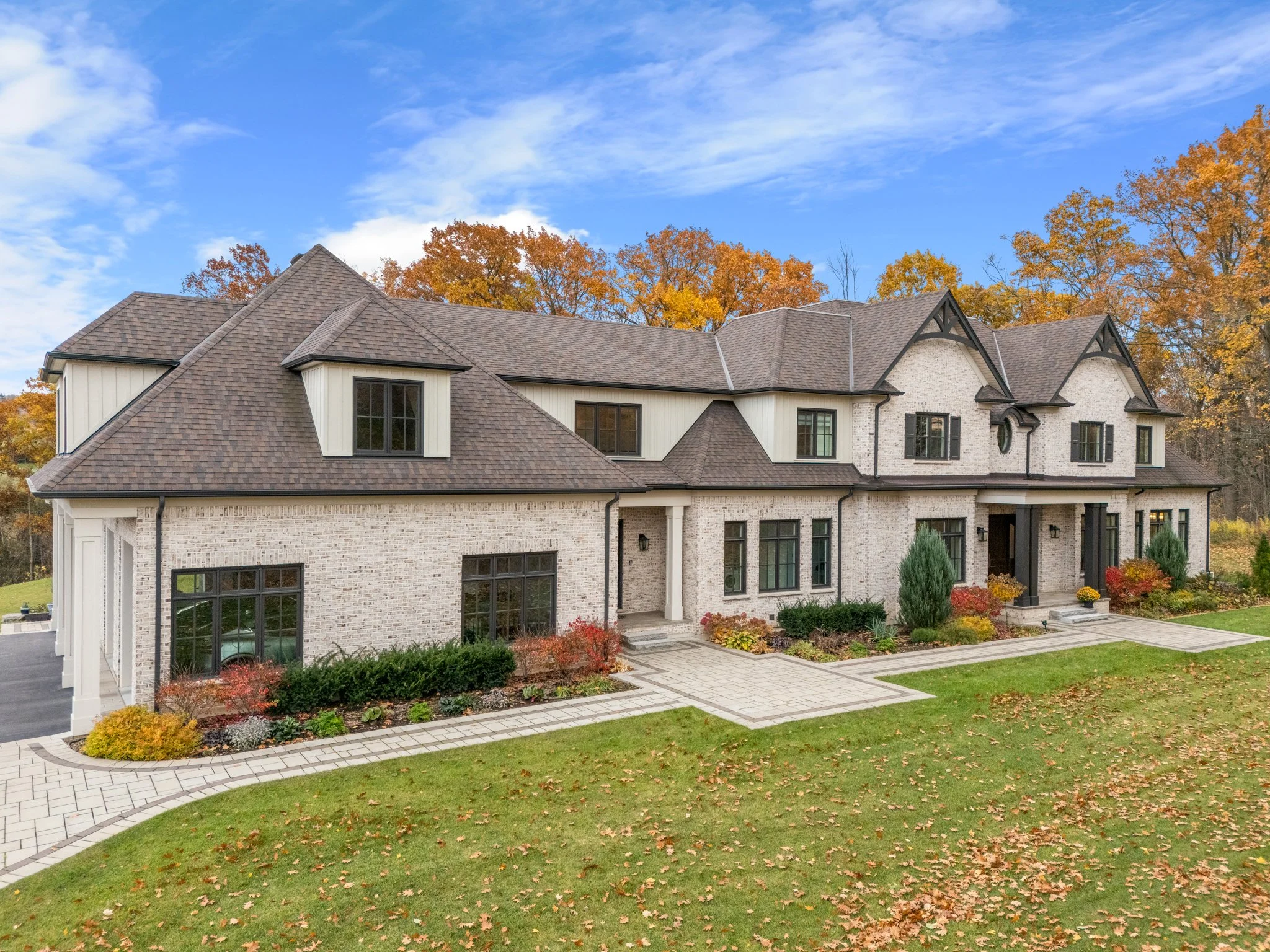 Large modern house with brick and siding exterior, multiple gable roofs, and a spacious front yard with a paved walkway, landscaped with bushes and trees during autumn.