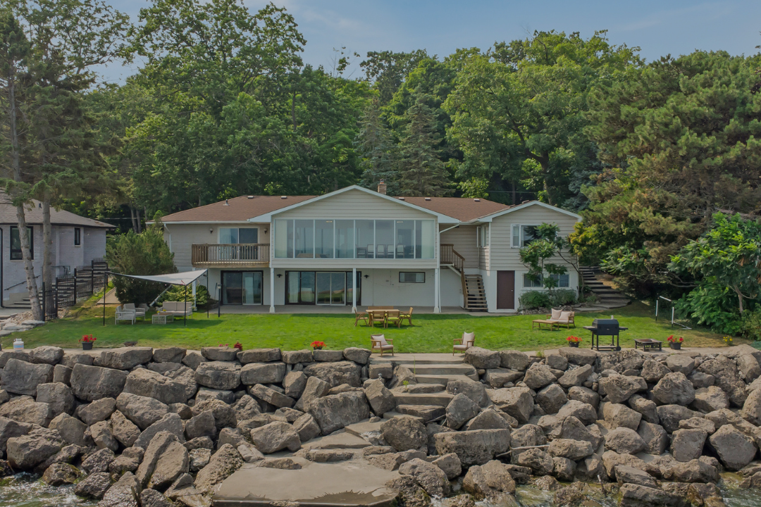 A house with a large deck and glass-enclosed porch faces a rocky shoreline with steps leading to the water. There are outdoor chairs, tables, and a barbecue grill in the yard, surrounded by green trees.