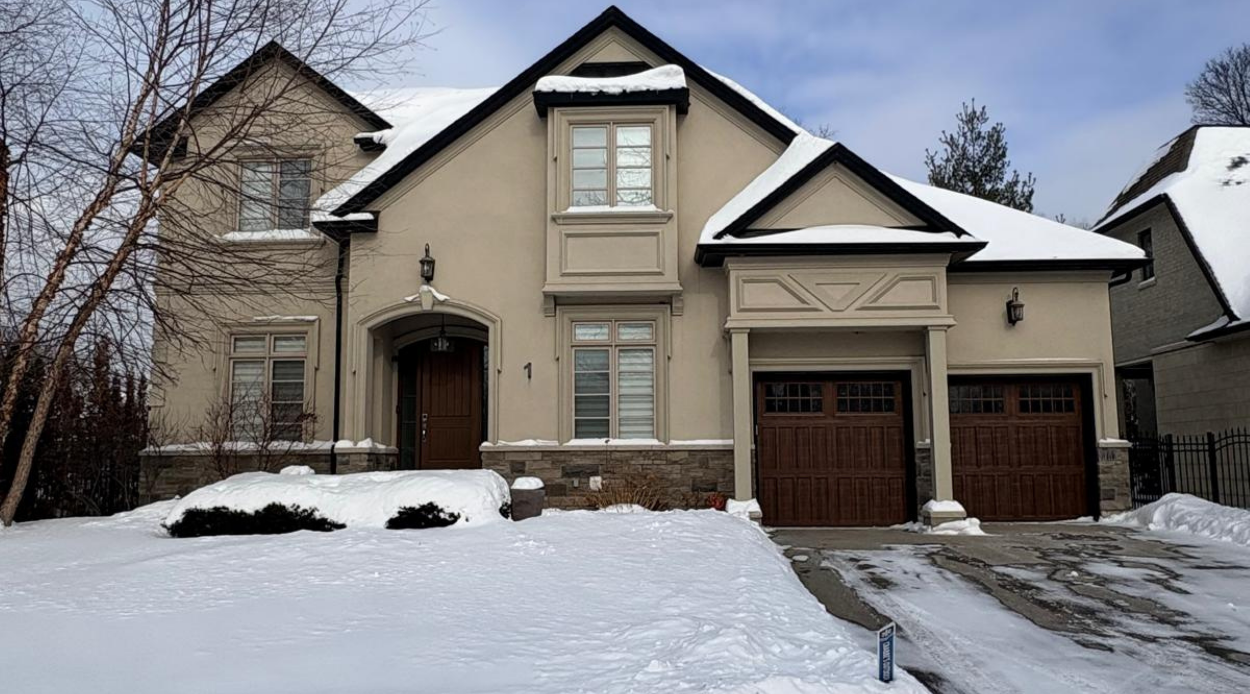 Front view of a two-story house with snow on the roof and yard, featuring a wooden front door, two garage doors, windows with shutters, and a stone foundation, amid leafless trees and neighboring houses.