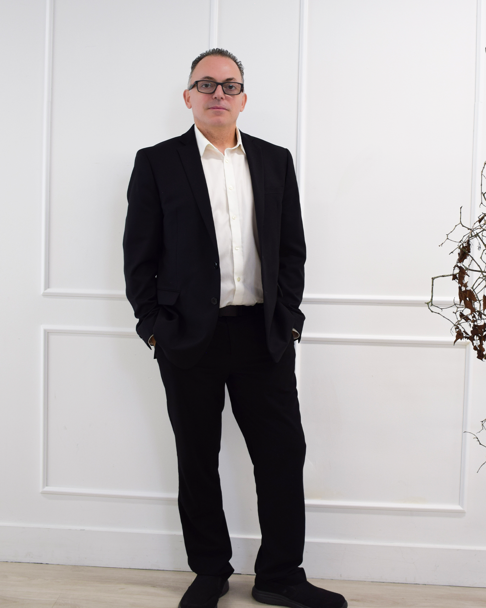 A man in a black suit and glasses standing in front of a plain white wall.