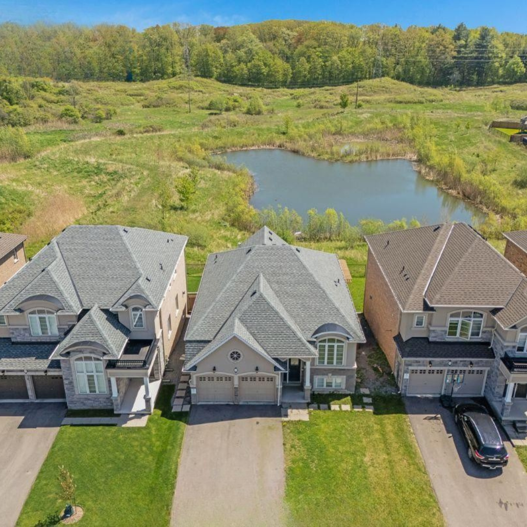 Aerial view of three suburban houses with gray and tan roofs, adjacent to a pond with lush greenery and trees in the background.