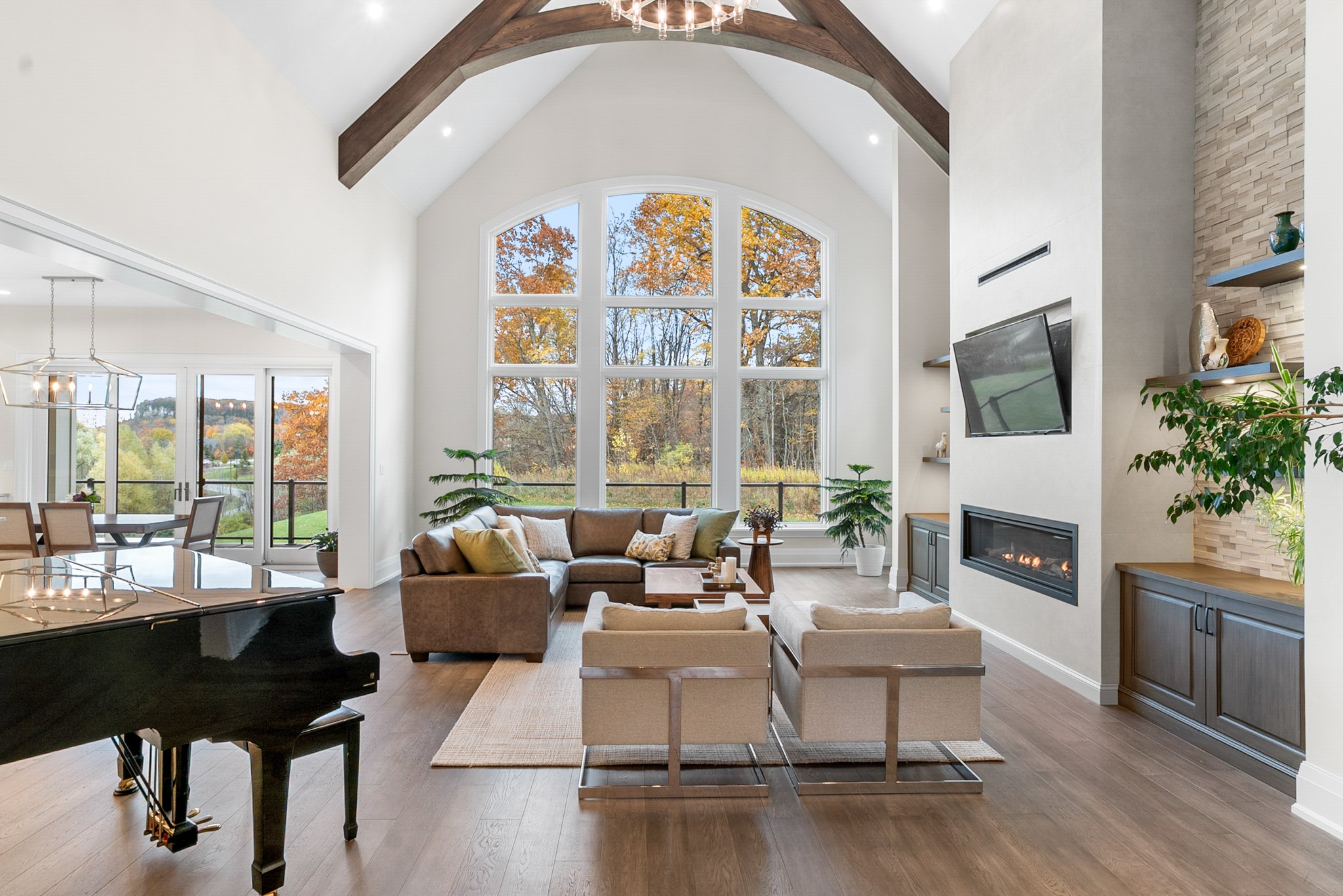 A spacious living room with a large arched window overlooking trees with autumn foliage, a brown sectional sofa with pillows, a fireplace with a mounted TV, a wooden coffee table, an area rug, and potted plants. There is a grand piano in the foreground and a dining area with a chandelier in the background.