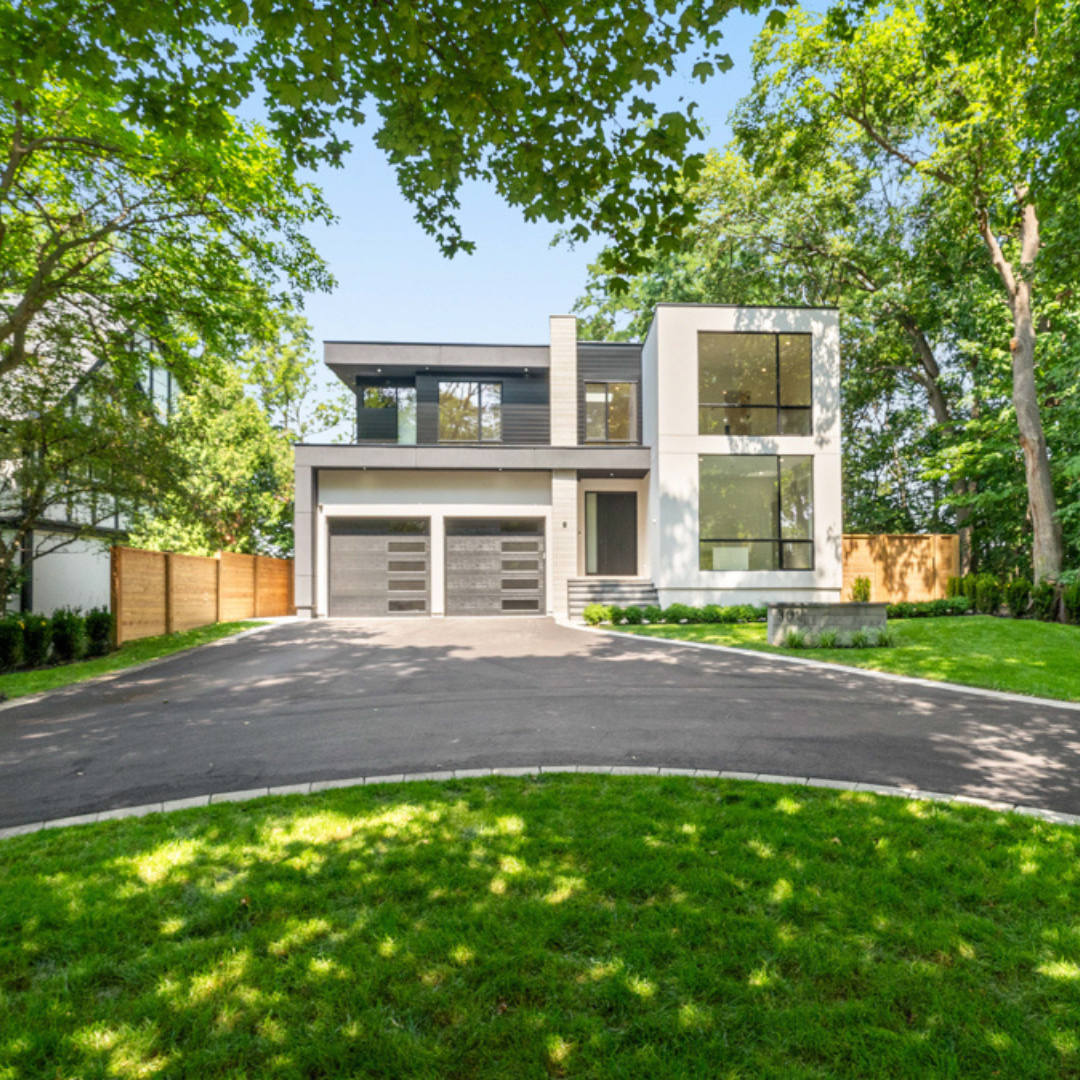 Modern two-story house with large windows, a dark driveway, and surrounding green trees and lawn.