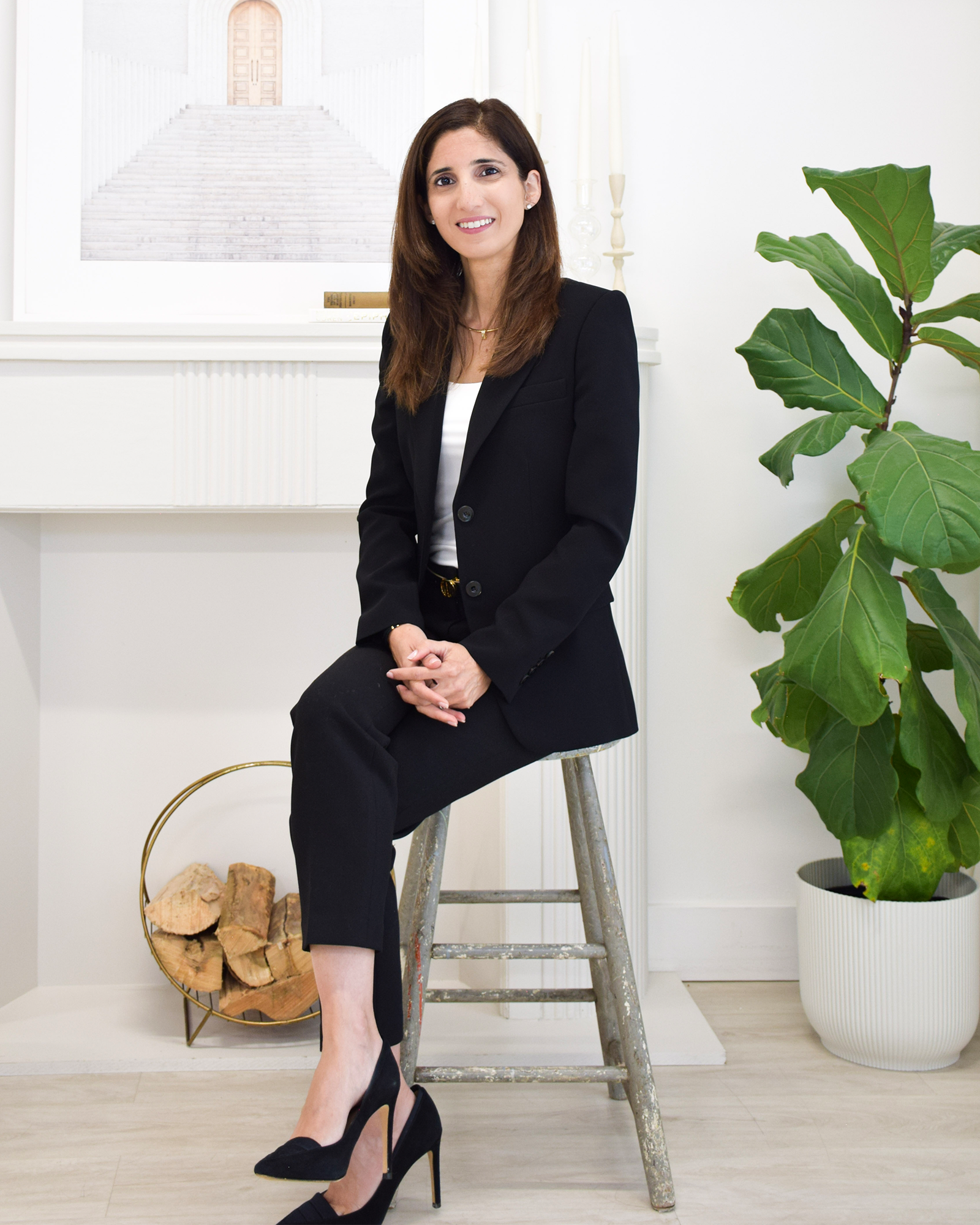 A woman in a black suit sitting on a wooden ladder in a bright room with a large green plant on her right and a framed art print of a staircase on her left, smiling at the camera.