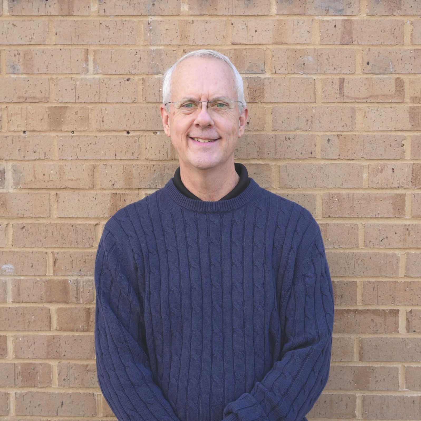 A smiling middle-aged man with glasses and short gray hair, wearing a navy blue cable-knit sweater, stands in front of a beige brick wall.