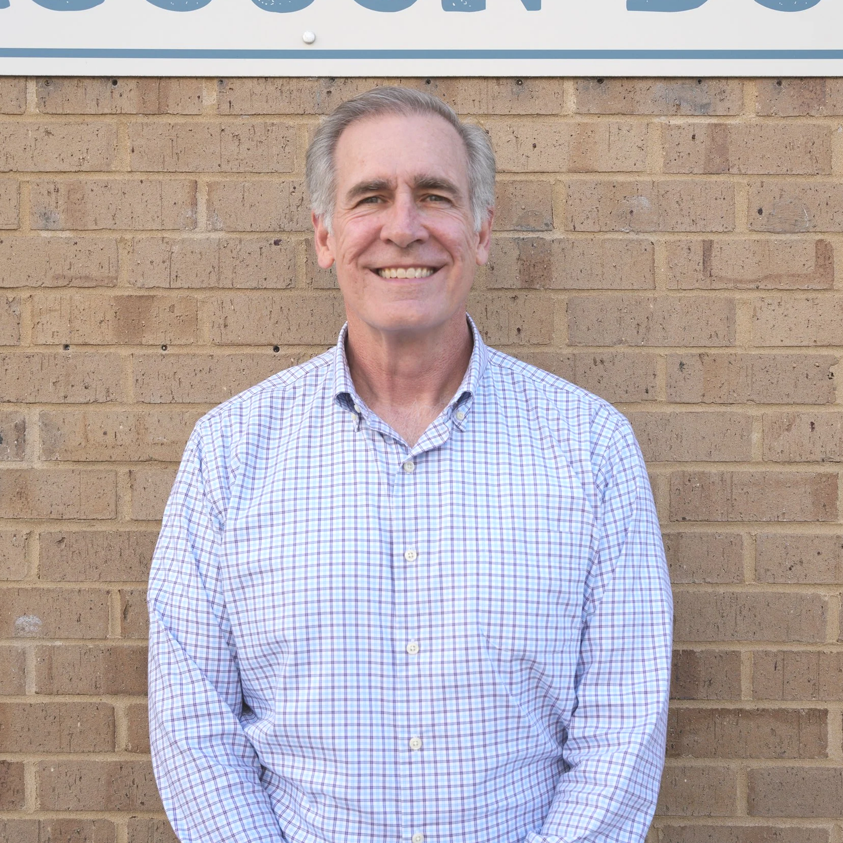 A smiling middle-aged man in a checkered shirt standing in front of a brick wall with part of a sign visible above his head.