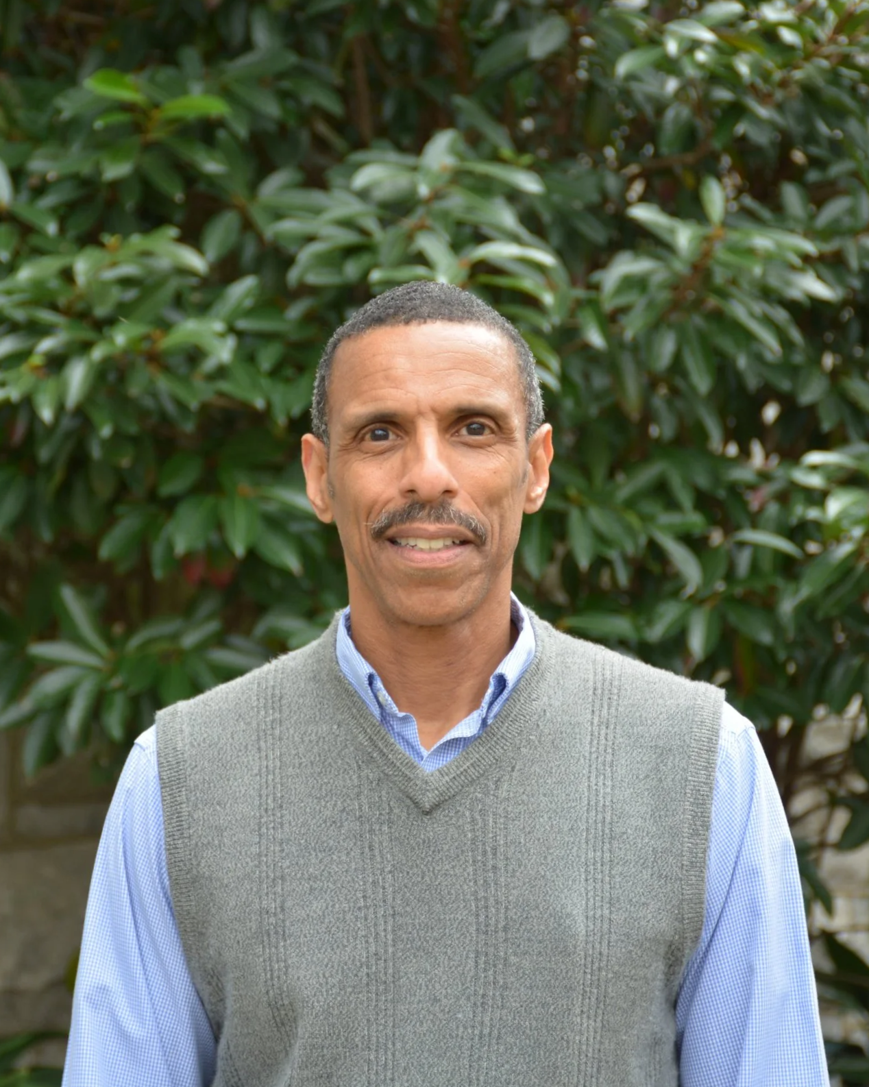 A man with short dark hair, a mustache, and light brown skin standing outdoors in front of green leafy bushes, wearing a light blue dress shirt and a gray sweater vest.