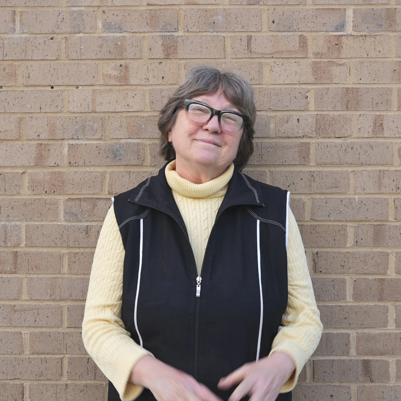 A woman with glasses and short brown hair stands in front of a brick wall, smiling softly with her eyes nearly closed. She is wearing a yellow turtleneck sweater and a black vest.