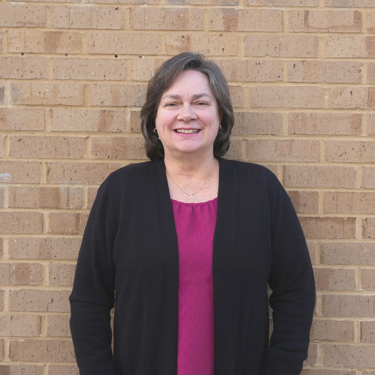A smiling middle-aged woman with shoulder-length gray hair, wearing a black cardigan over a magenta top, standing in front of a brick wall.