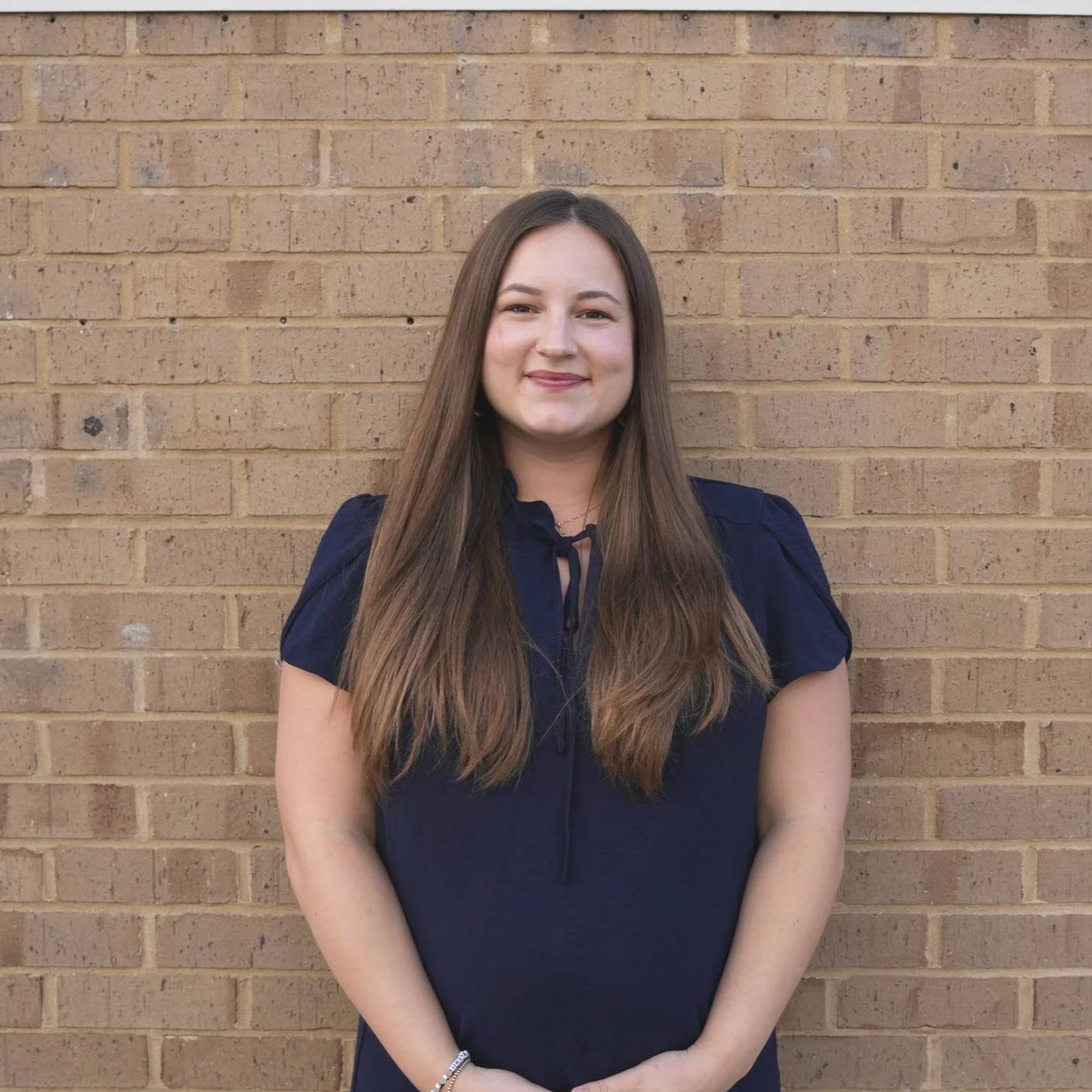 A young woman with long brown hair, smiling, standing in front of a brick wall, wearing a navy blue short-sleeve top.