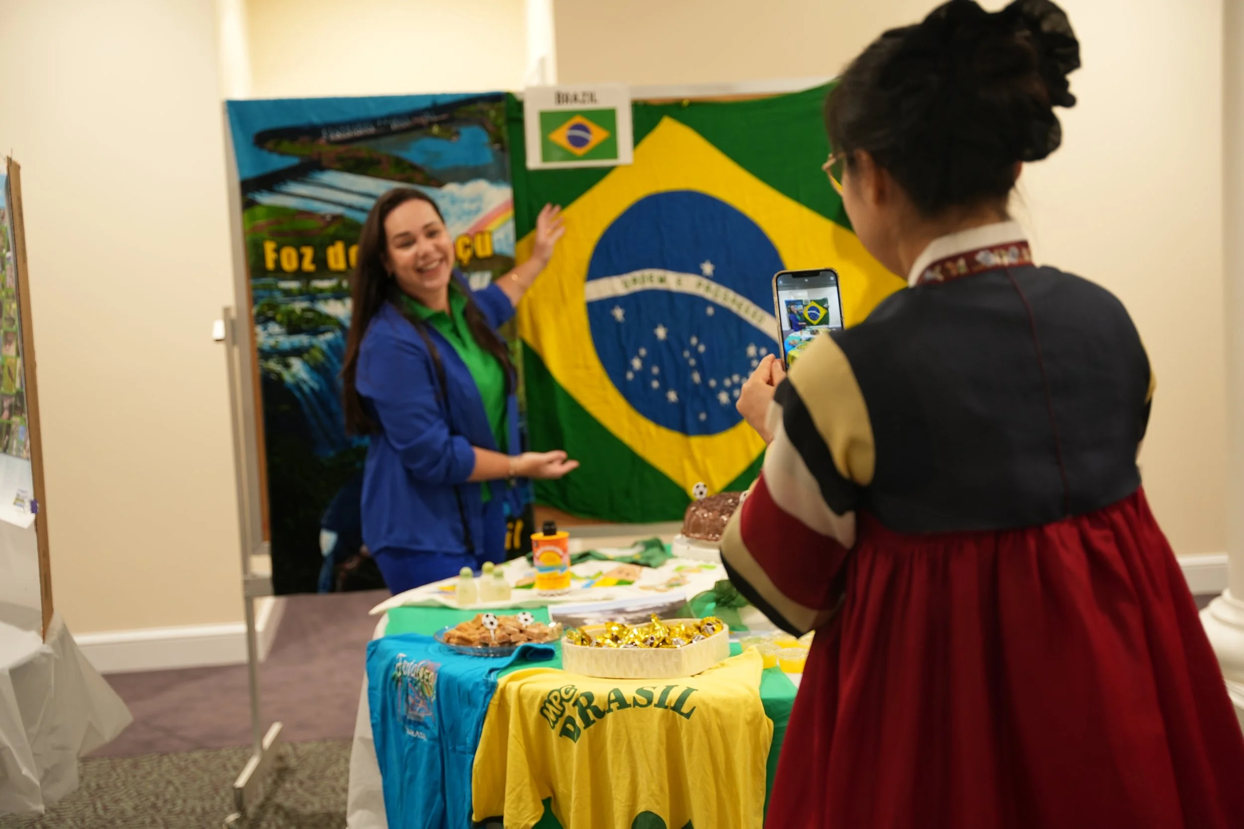 Two women, one smiling and wearing a blue jacket, stand in front of a display with the Brazilian flag. One woman is taking a photo of the other with her phone. The table nearby has various items, including a yellow shirt with green and yellow text and a small soccer ball decoration.