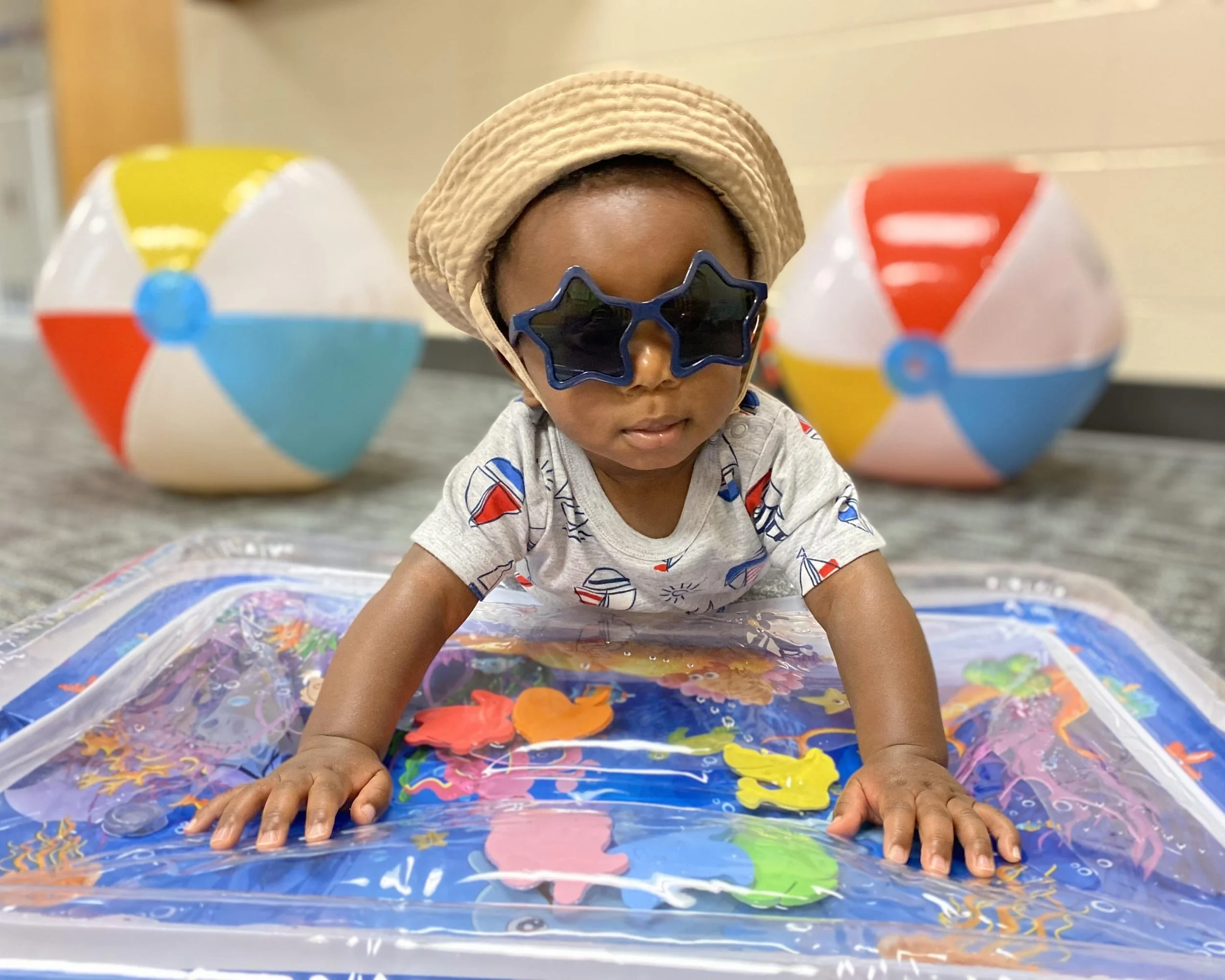 A young child wearing star-shaped sunglasses, a sunhat, and a surf-themed shirt, crawling on a water playmat with colorful fish and sea creatures, with beach balls in the background.