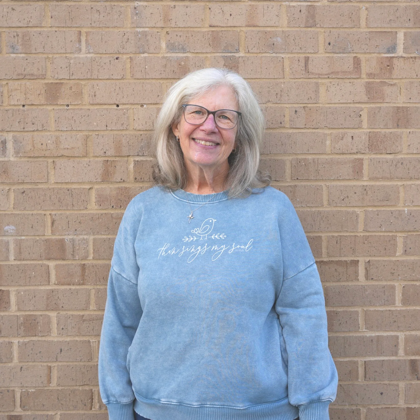 Smiling elderly woman with glasses and gray hair, wearing a light blue sweatshirt with a bird and the phrase "he who sings my soul," standing in front of a brick wall.