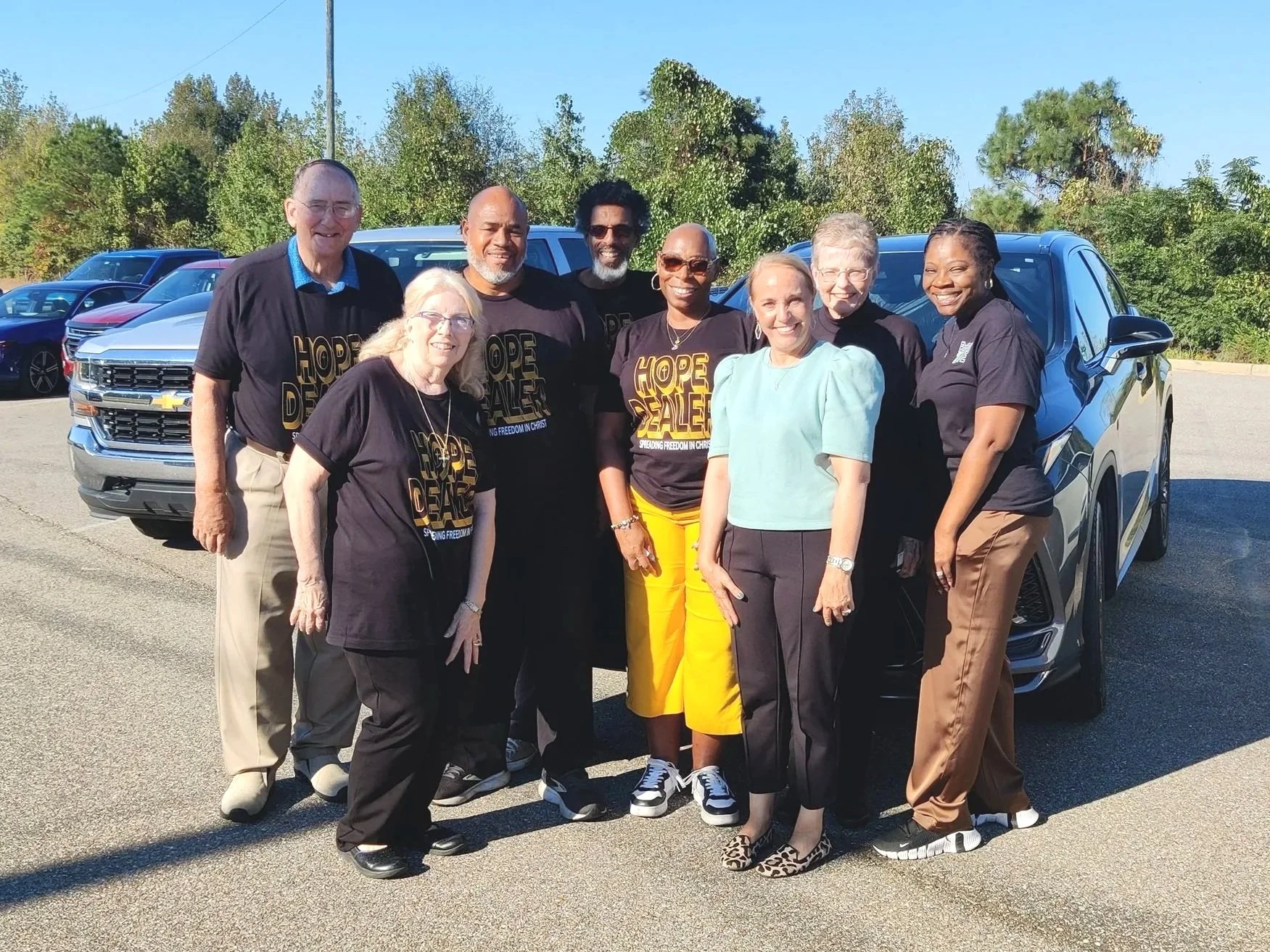 A group of diverse people standing outdoors on a sunny day, smiling at the camera, in a parking lot with trees and cars in the background.