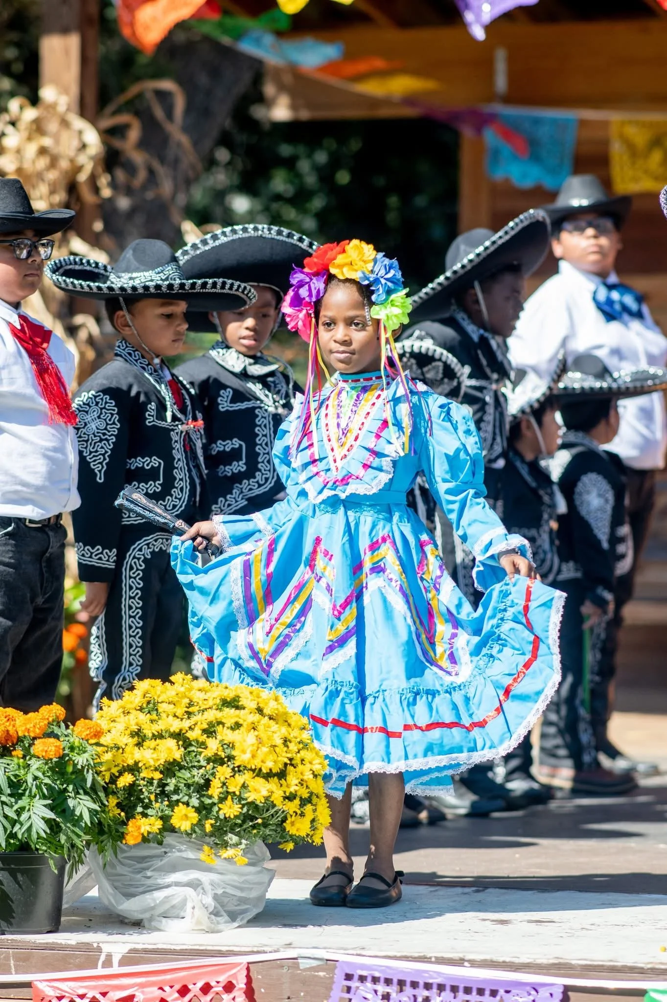 A group of children dressed in traditional Mexican mariachi attire, with one girl in the center wearing a bright blue dress with colorful embroidery and a large flower crown, participating in a cultural celebration or performance outdoors.