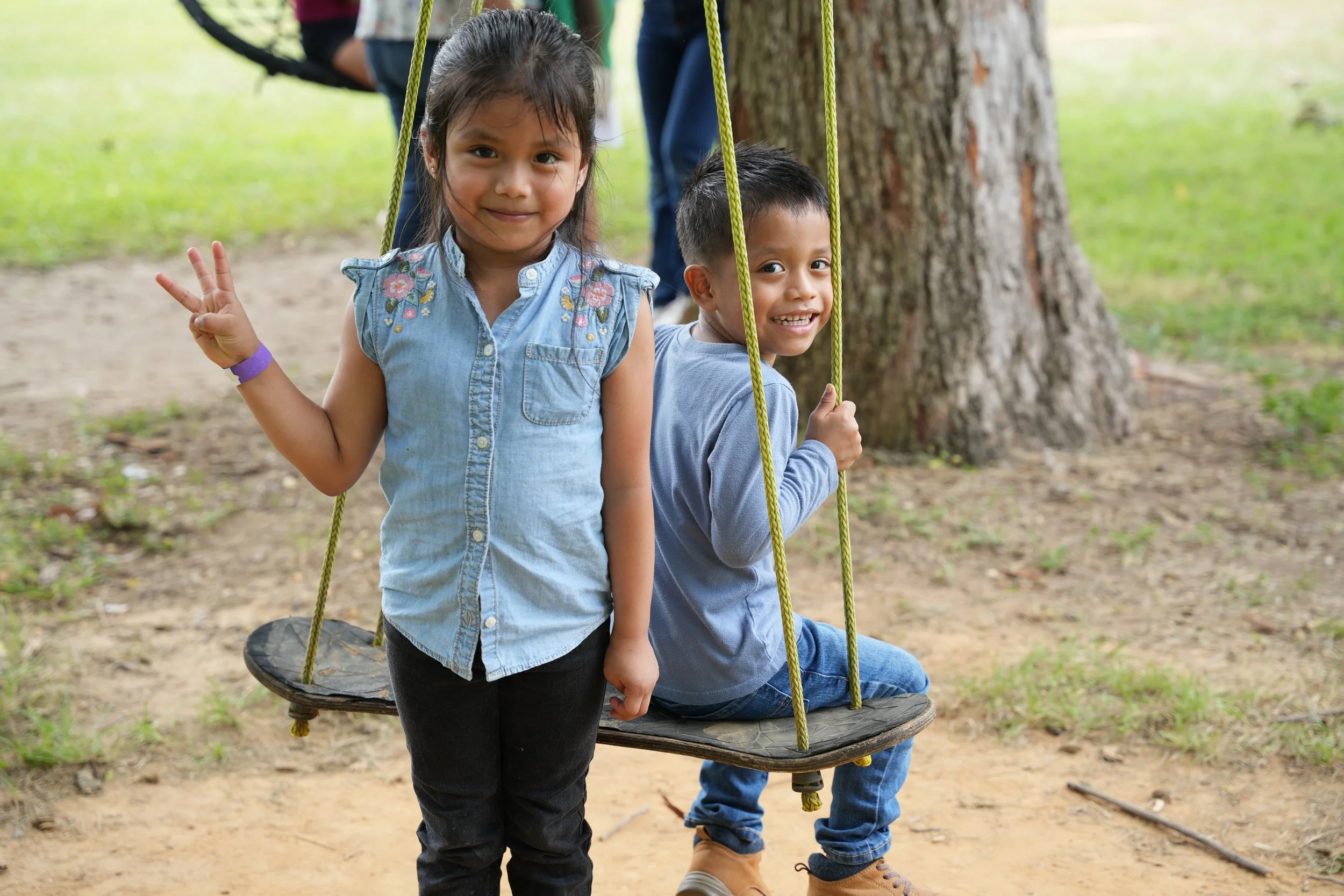 Two kids playing at a park; a girl standing in front making a peace sign, and a boy sitting on a swinging seat attached to a tree trunk, both smiling.