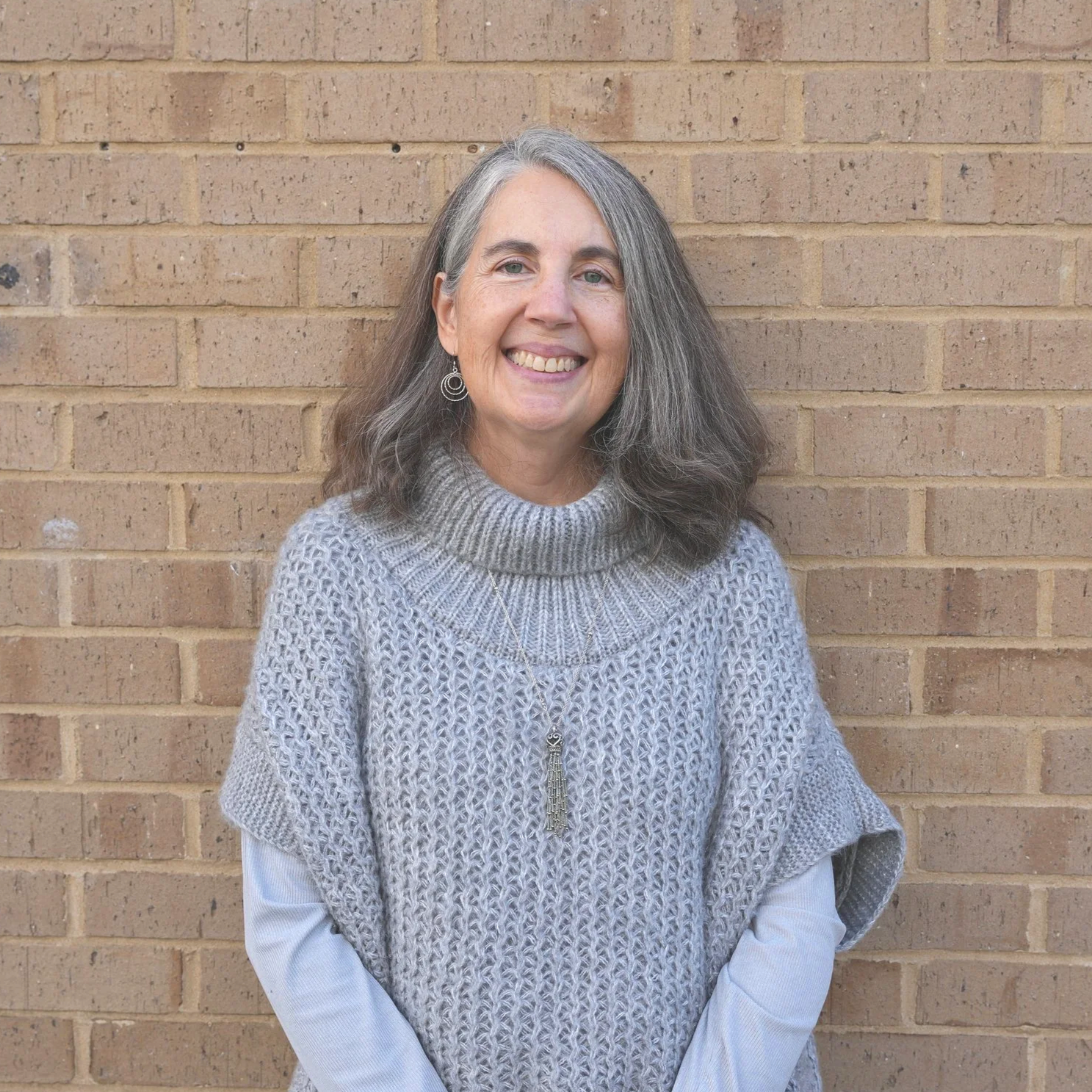 A smiling woman with gray hair, wearing a cozy gray knit top layered over a white long-sleeve shirt, standing in front of a brick wall.