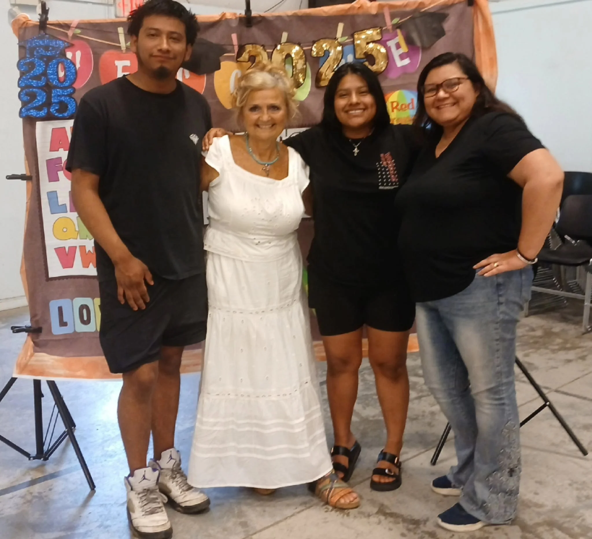 Four people standing together in front of a colorful decorated backdrop for a birthday celebration. The backdrop displays the numbers '2025' and other colorful letters. The group includes a young man, an older woman in a white dress, and two women in black shirts and jeans, all smiling and posing for the photo.