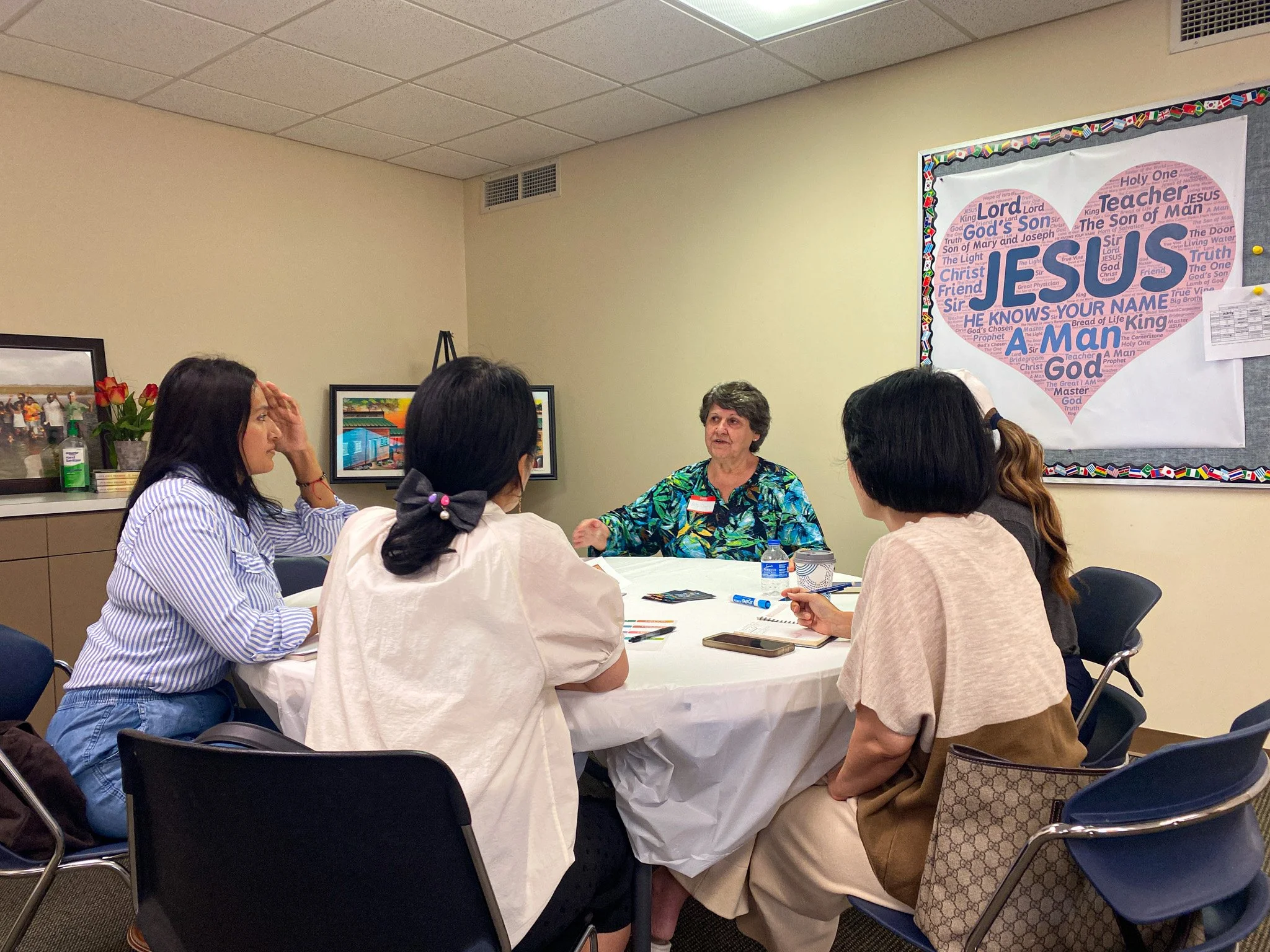 A group of women sitting around a round table in a meeting room, listening to an older woman speaking. The room has artwork on the walls, including a large poster with a heart shape made of words related to Jesus and Christianity.