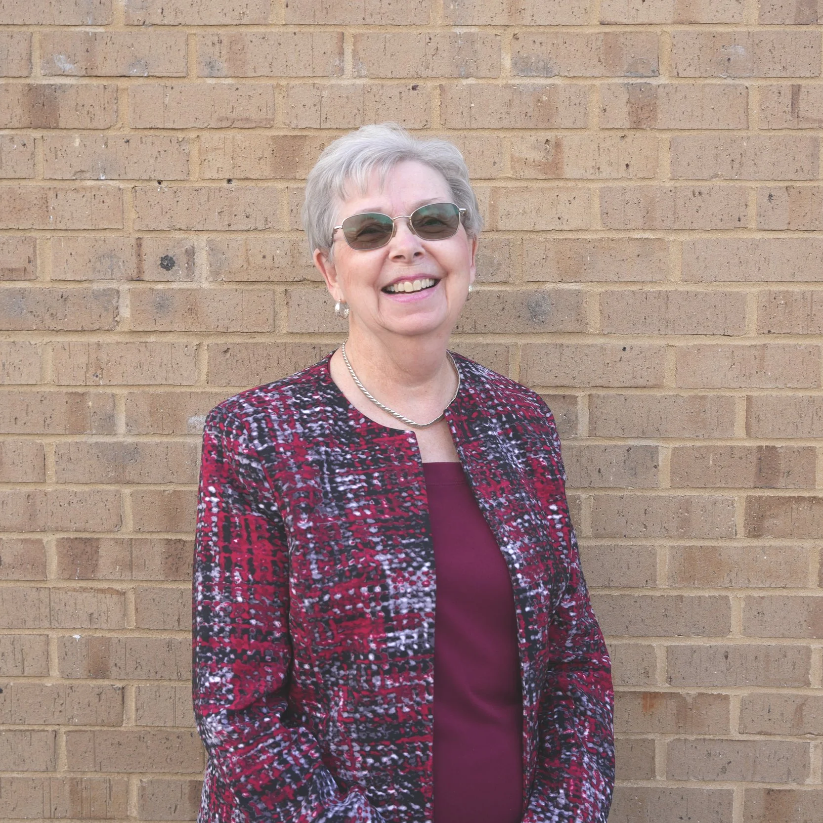Smiling elderly woman with short gray hair, sunglasses, earrings, a pearl necklace, and a patterned red and black blazer standing against a brick wall.