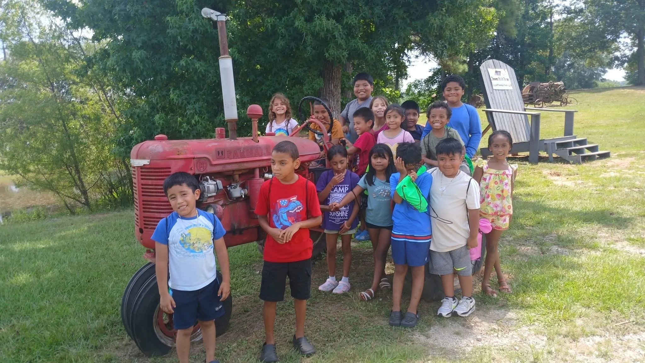 A group of children gathered outdoors on a grassy area with trees in the background, posing next to a vintage red tractor and a wooden sign. The children are smiling, dressed casually, and some are holding water bottles or backpacks.