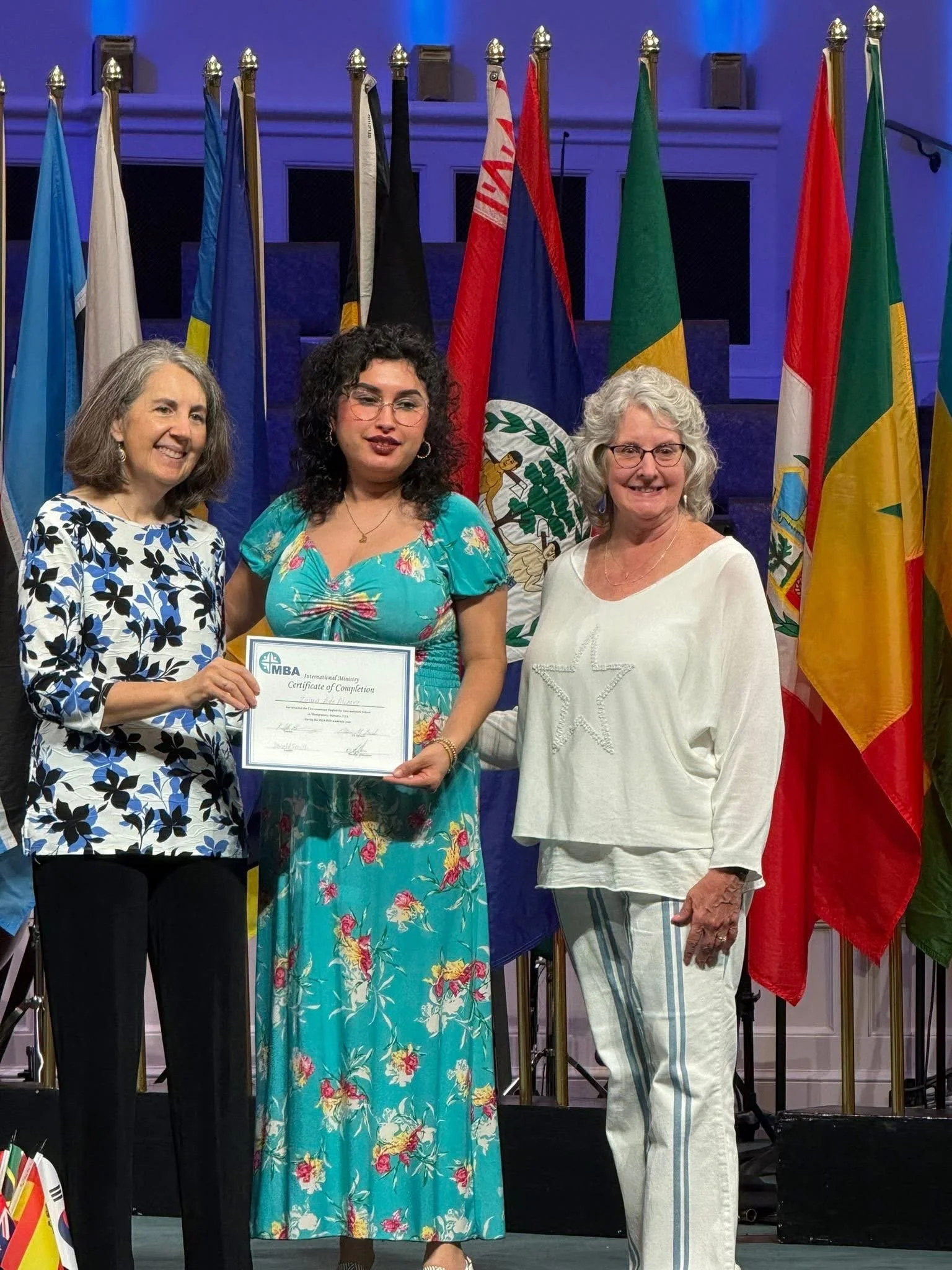 Three women standing on a stage with international flags in the background. One woman holds a certificate of completion, and they are smiling at the camera.