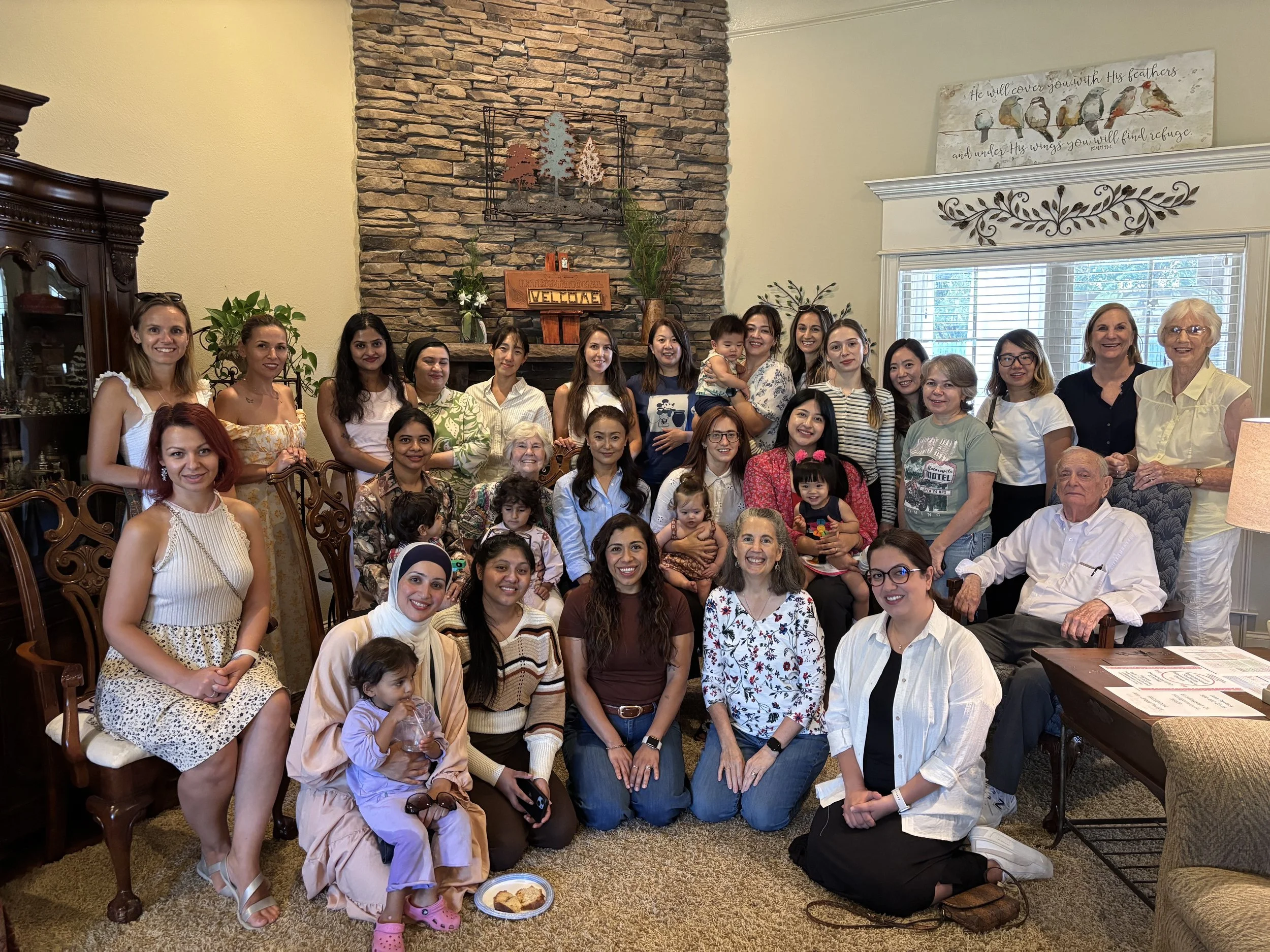 A large group of diverse women and children gathered in a living room for a family or community gathering, smiling at the camera.