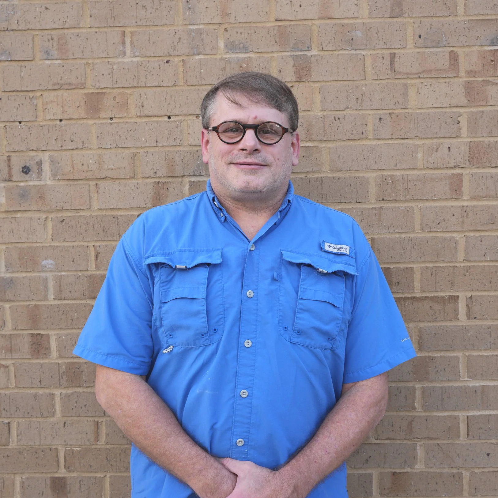 A man with glasses and short hair standing in front of a brick wall, wearing a blue short-sleeved button-up shirt.