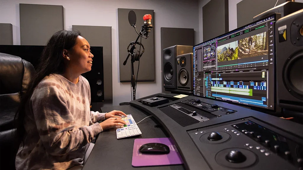 A woman working in an audio and video editing studio with a large monitor displaying editing software, audio speakers, a microphone, and control panels.