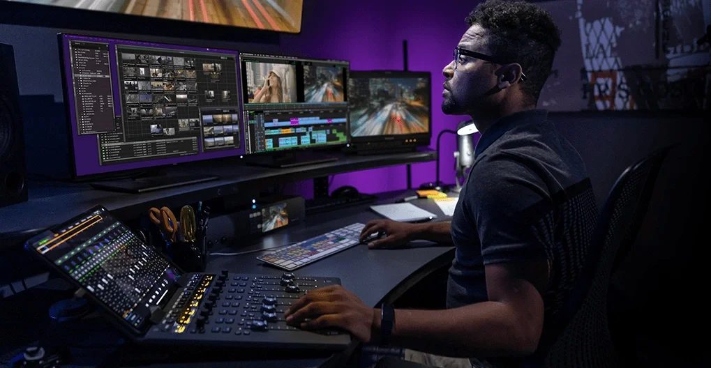 A person working at a multi-monitor editing station with video editing software, audio mixing console, and professional audio equipment in a dark room with purple lighting.