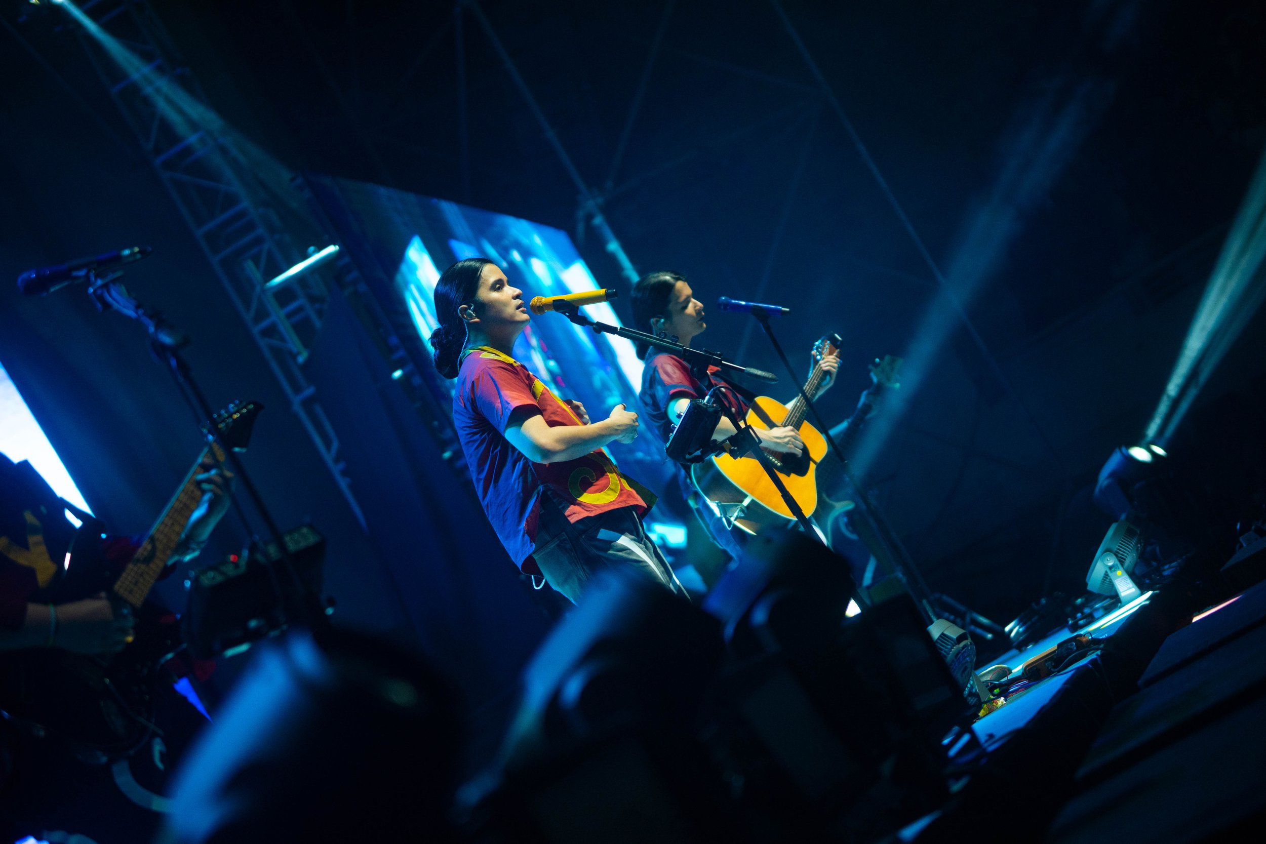 Two singers and a guitarist performing on stage with blue lighting and stage equipment.