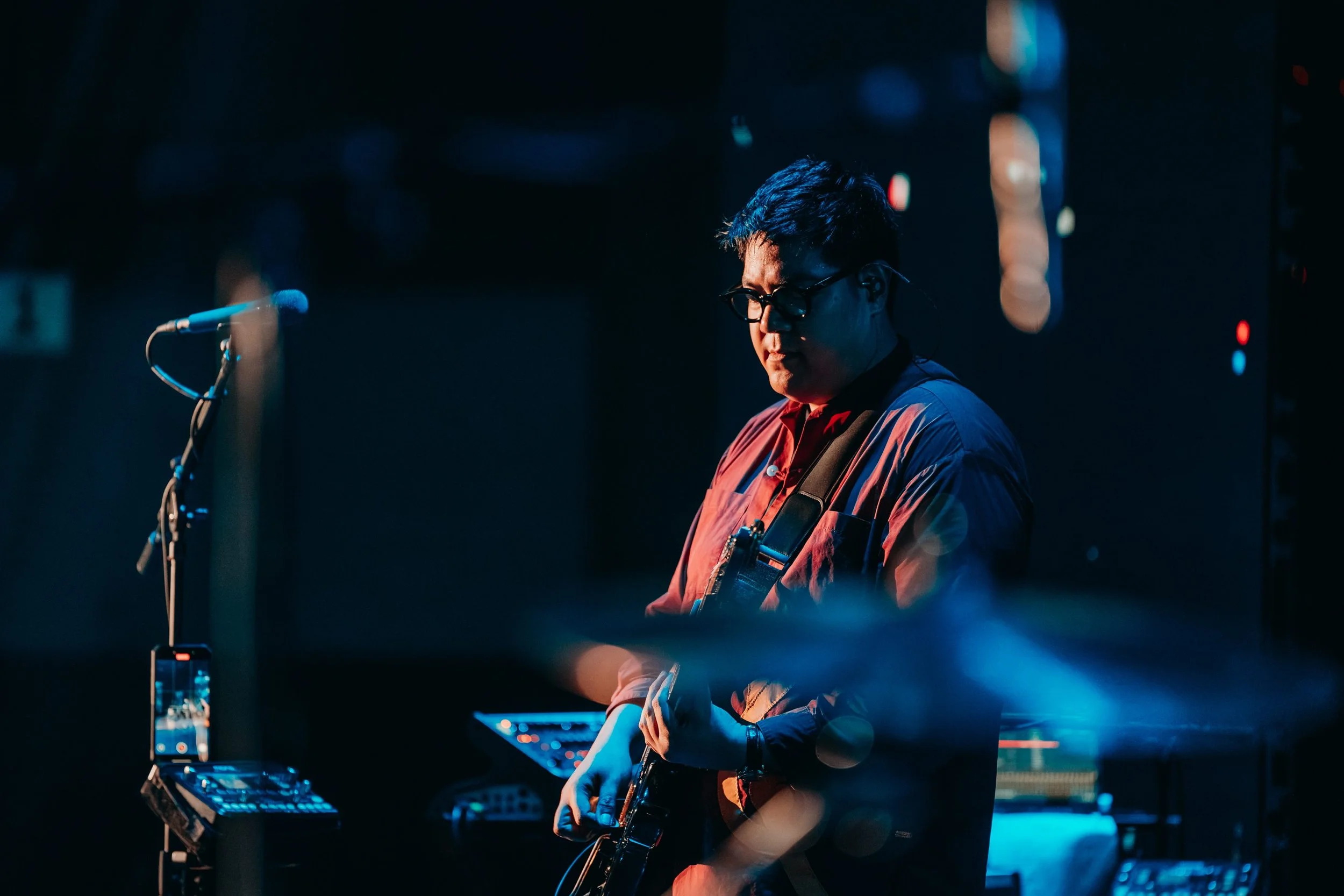 A musician playing an electric guitar on stage, wearing glasses and a dark shirt, with music equipment around him, illuminated by stage lighting.