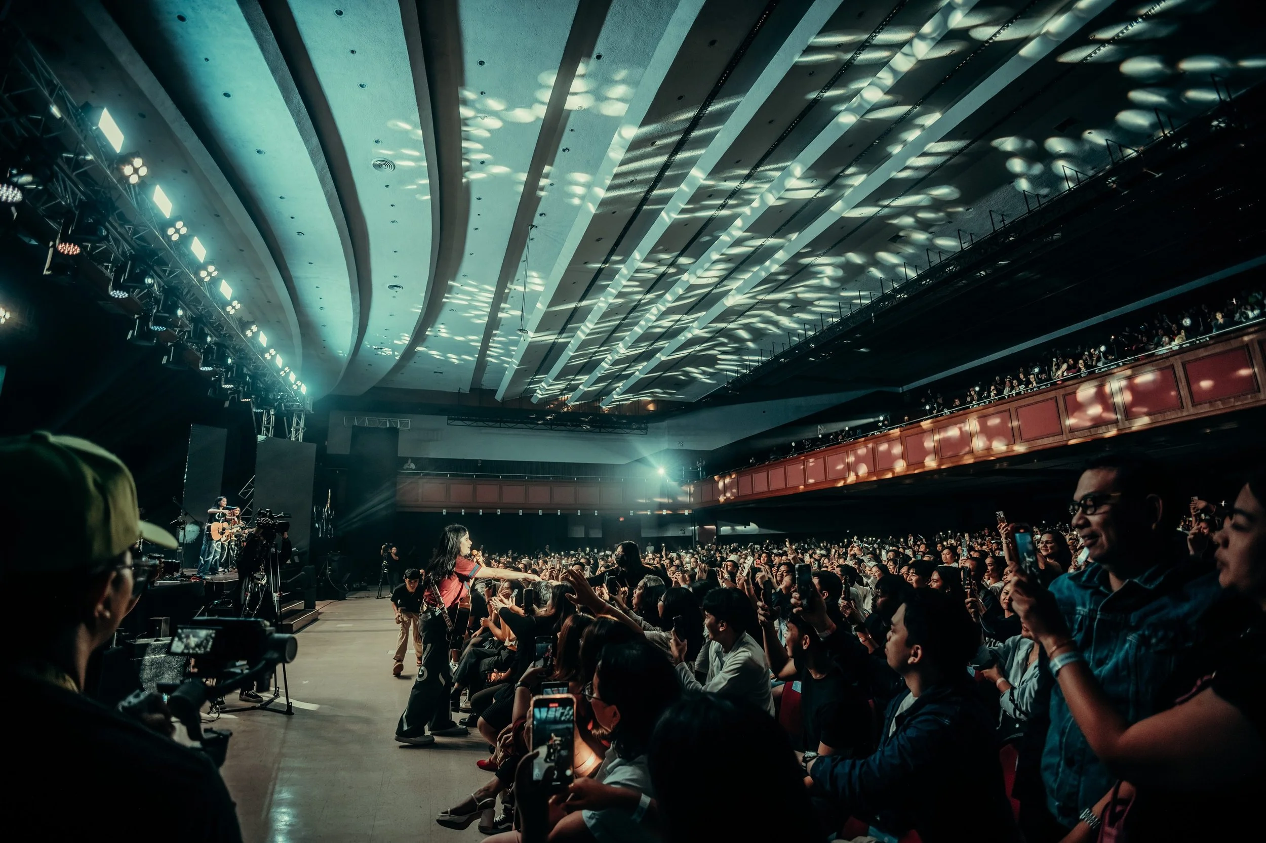 Crowd attending live concert or performance in an indoor theater with stage lighting and musicians playing on stage.