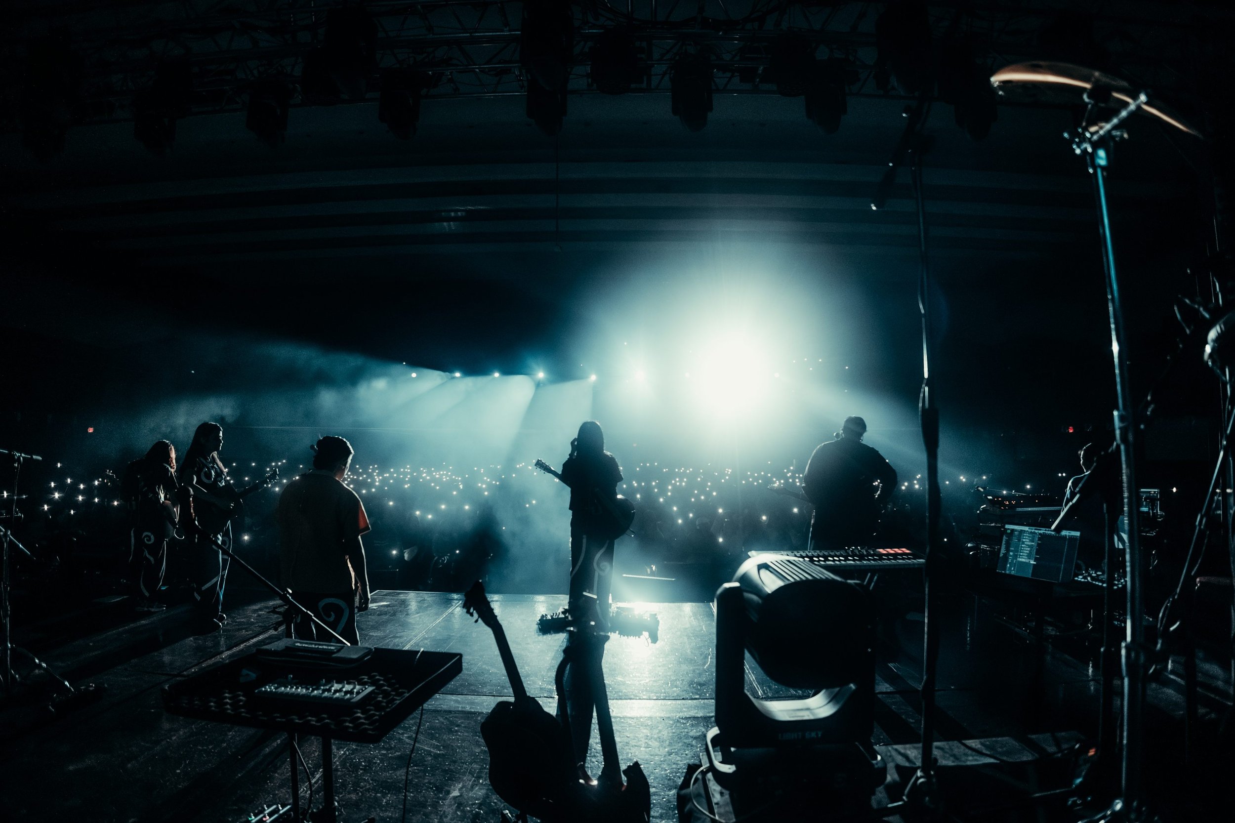 Silhouettes of a band performing on stage with bright lights and an audience holding up phones in the background.