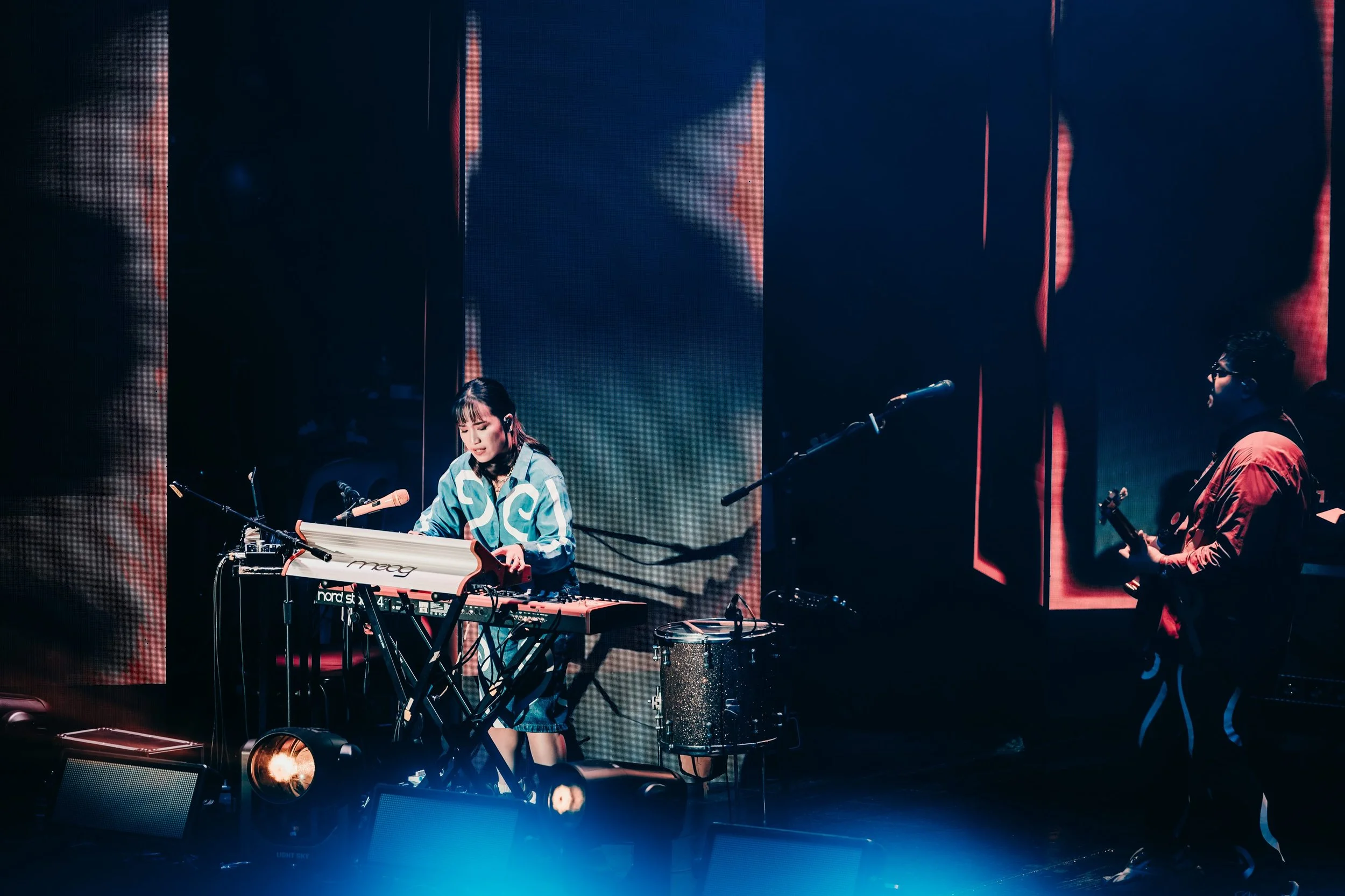 A female musician playing keyboard on stage with a male guitarist beside her, with stage lighting and screen background.