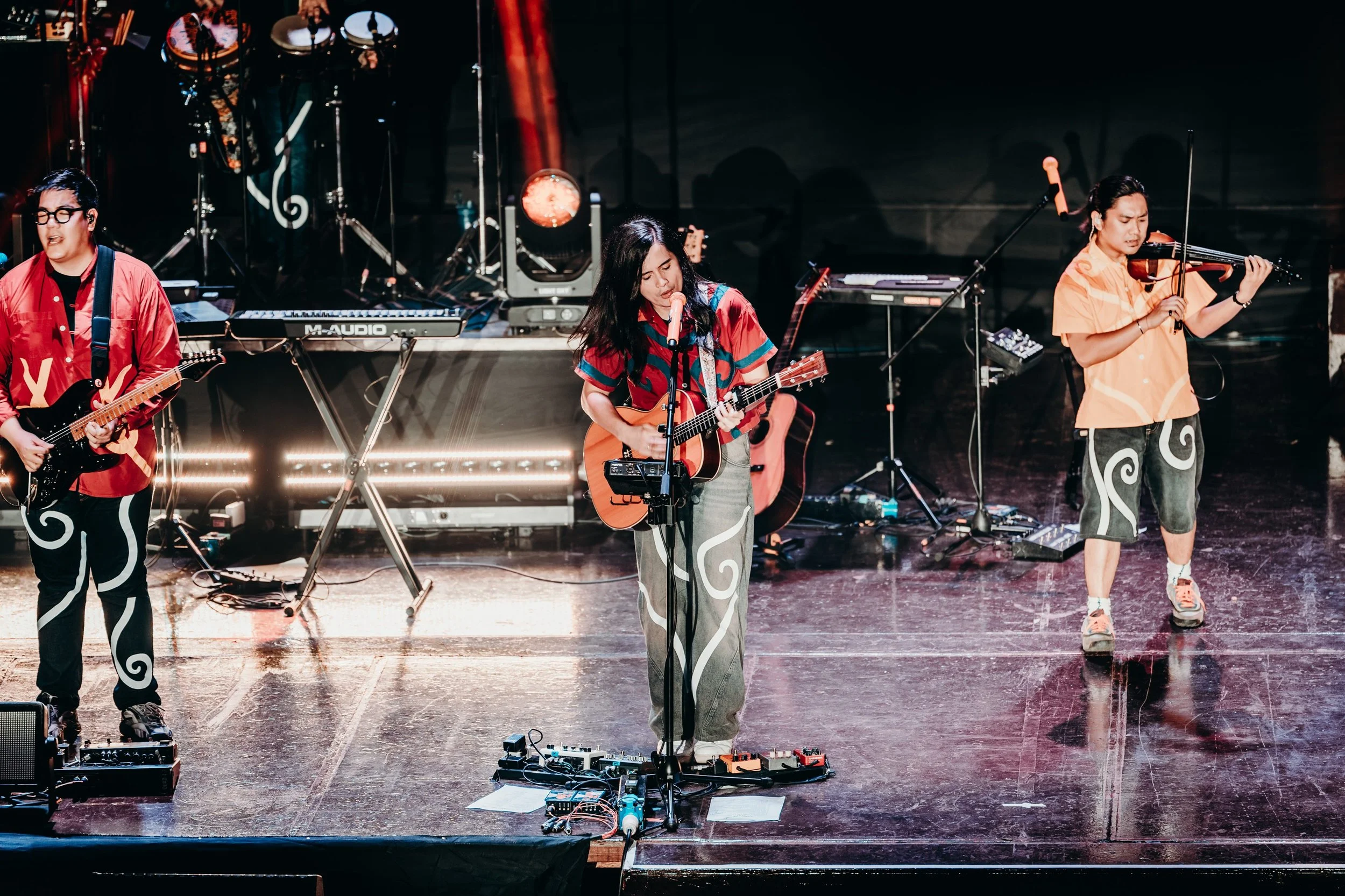 Three musicians performing on stage with guitars, keyboard, and violin in a dimly lit concert setting.