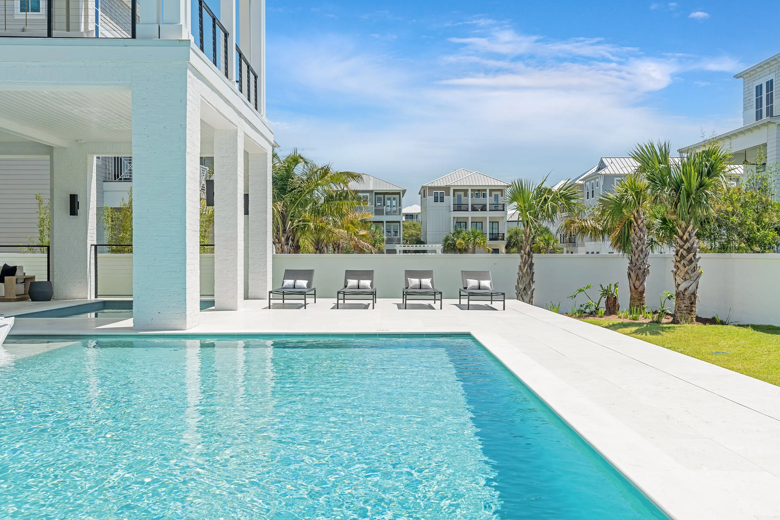 A modern outdoor swimming pool area with white tiled deck, four black lounge chairs with white pillows - Real estate photography in 30A, Destin, and Panama City Beach by Matt Jozwiak