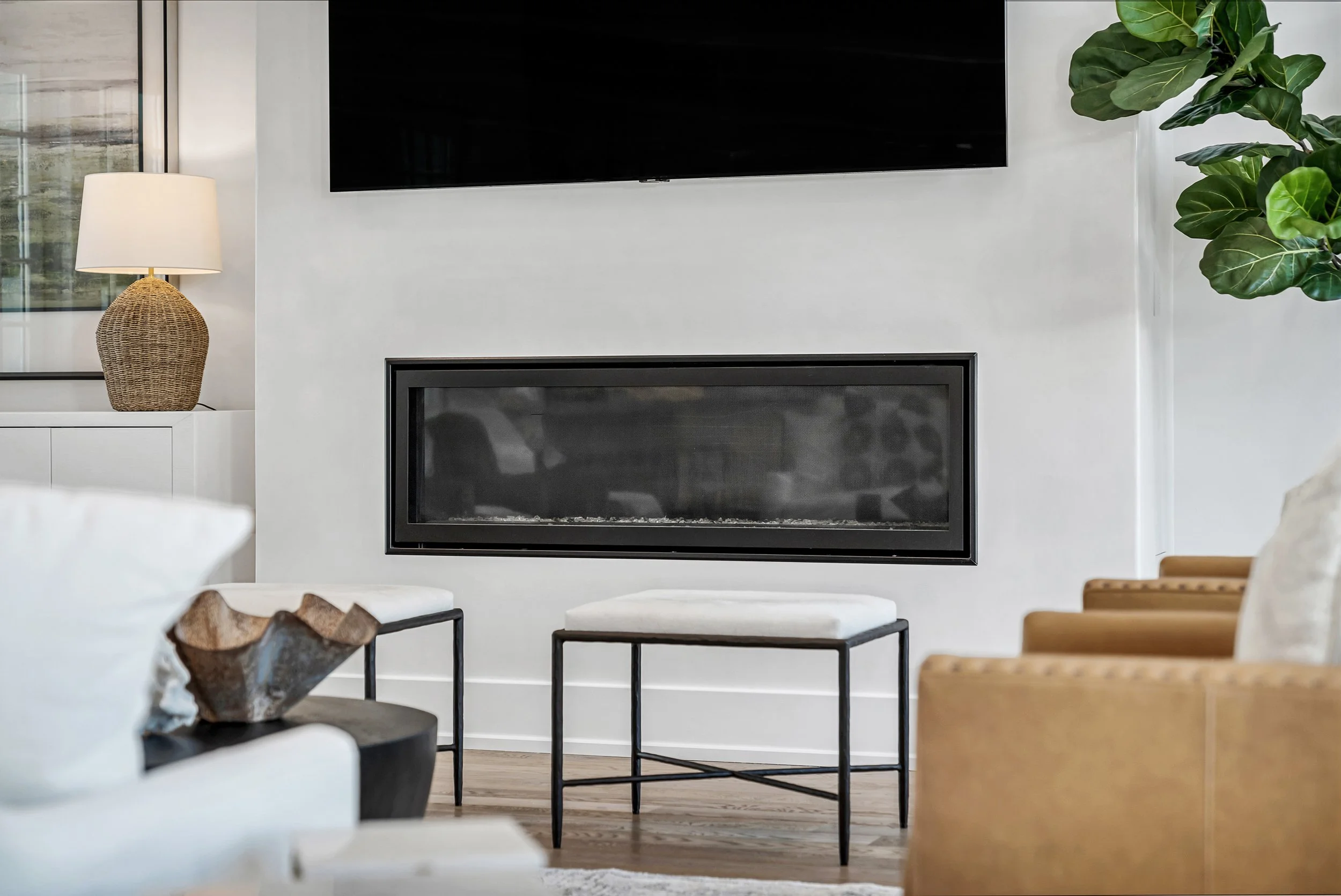 Living room with a fireplace, wall-mounted TV, a table with logs, a white armchair, a beige armchair, a white cushion, a side table, a large green plant, and a lamp with a woven base - Real estate photography in 30A, Destin, and Panama City Beach by
