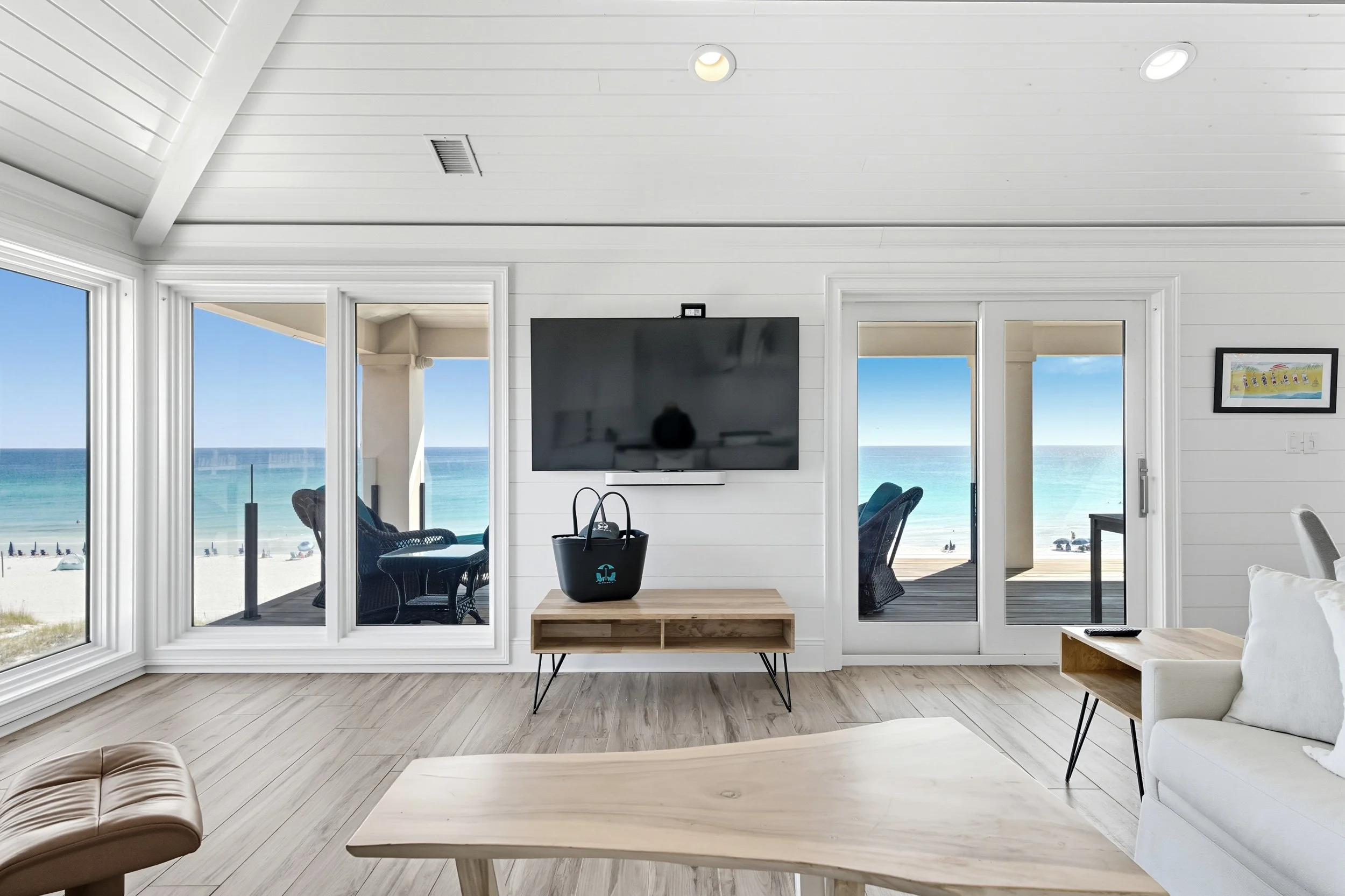 Living room with large windows and ocean view, white walls, light wood flooring, mounted flat-screen TV, small wooden table, and outdoor patio furniture visible through glass doors - Real estate photography in 30A, Destin, and Panama City Beach by Ma