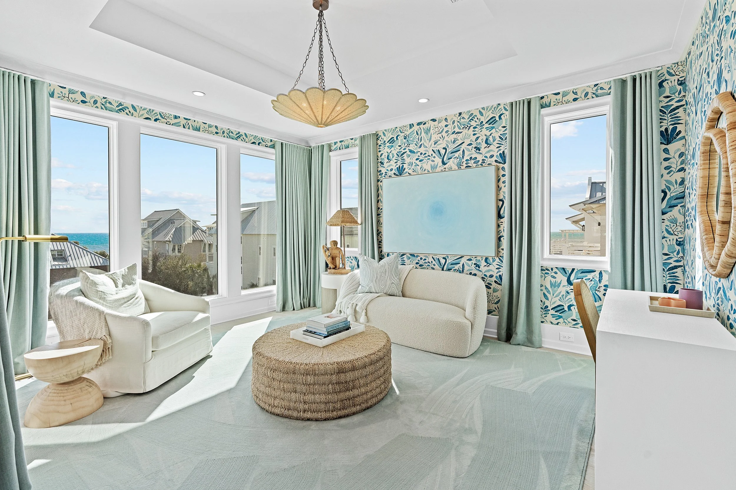 Bright coastal living room with large windows, ocean view, white sofa, armchair, round woven coffee table, blue patterned wallpaper, green curtains, and minimalist decor - Real estate photography in 30A, Destin, and Panama City Beach by Matt Jozwiak