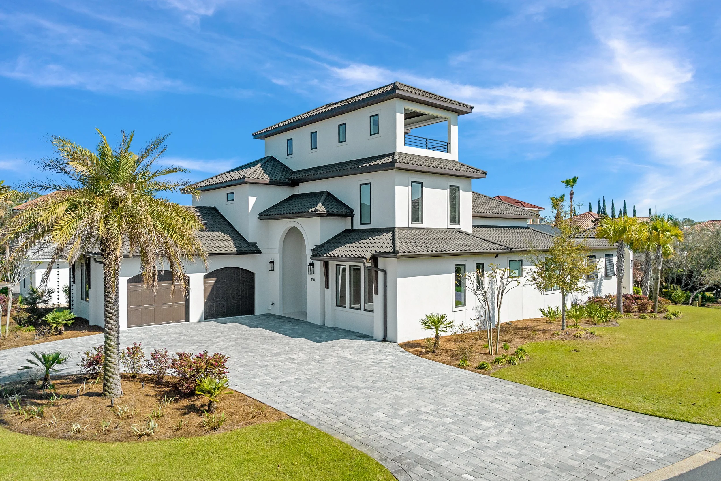 Large modern white house with multiple levels, grey tile roof, and attached garage, surrounded by landscaped yard with palm trees and a paved driveway, under a clear blue sky - Real estate photography in 30A, Destin, and Panama City Beach by Matt Joz