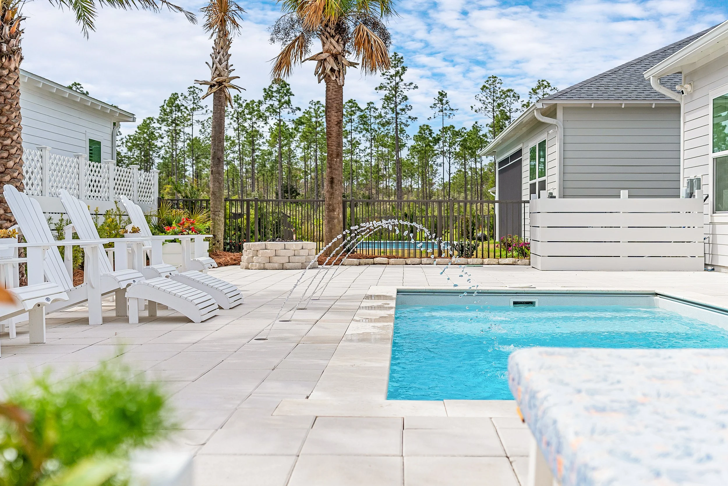 A backyard patio with a swimming pool, white lounge chairs, palm trees, and a black fence with a view of a backyard and trees - Real estate photography in 30A, Destin, and Panama City Beach by Matt Jozwiak