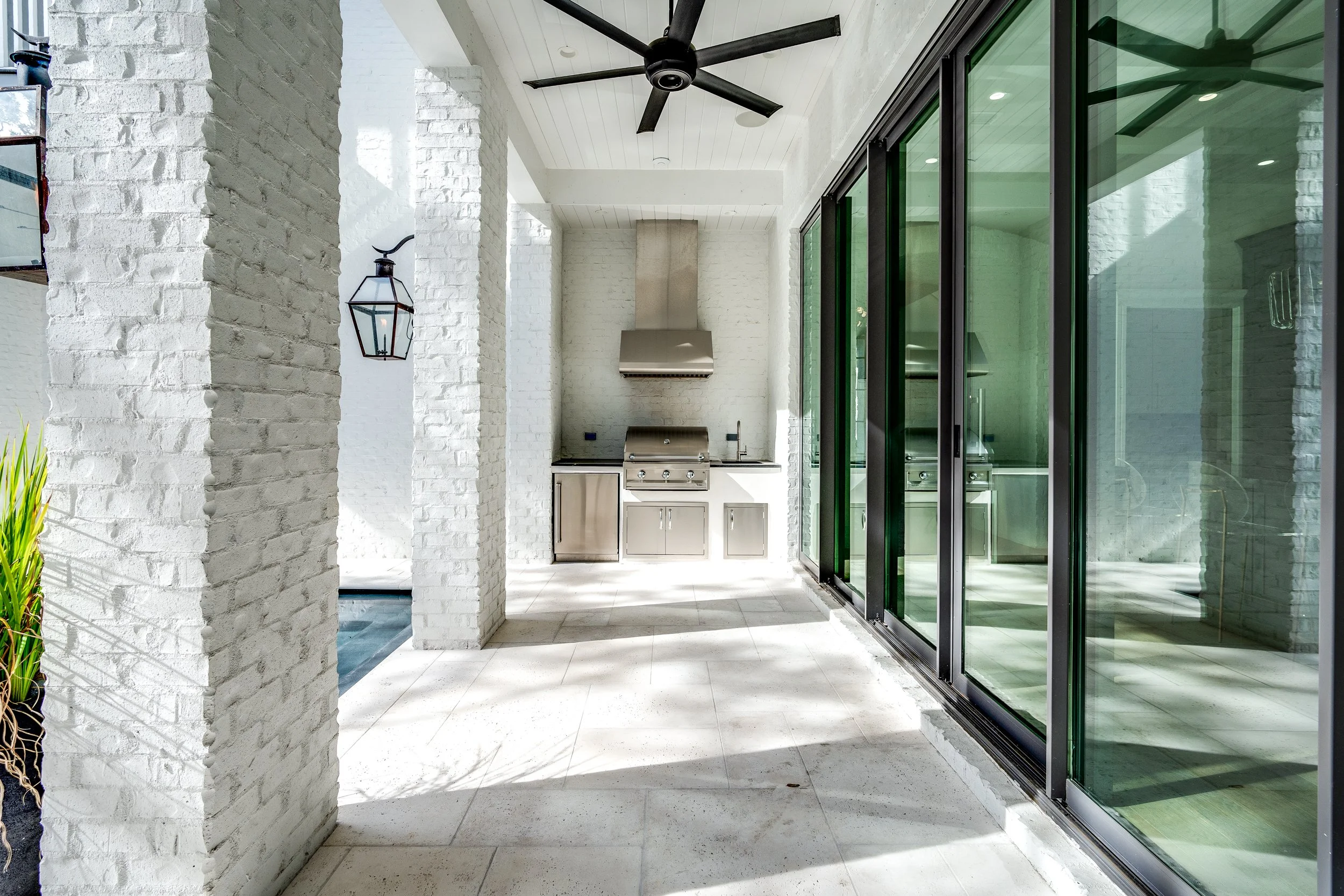 Outdoor kitchen with stainless steel grill, white brick walls, large glass sliding doors, ceiling fan, and lantern-style wall light - Real estate photography in 30A, Destin, and Panama City Beach by Matt Jozwiak