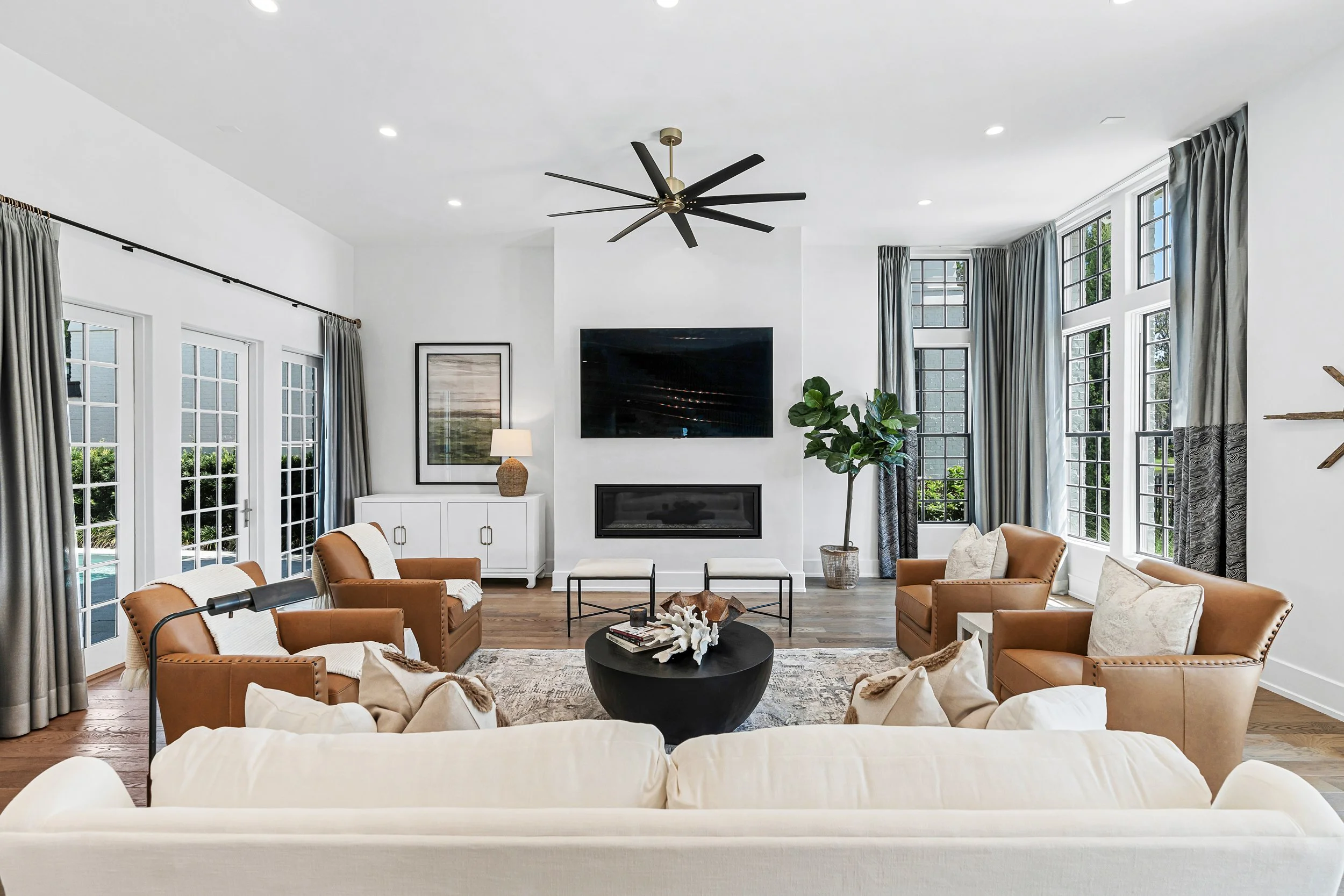 Living room with white walls, large windows with gray curtains, and a central black ceiling fan. There are six armchairs and a white sofa arranged around a black coffee table. - Real estate photography in 30A, Destin, and Panama City Beach by Matt J