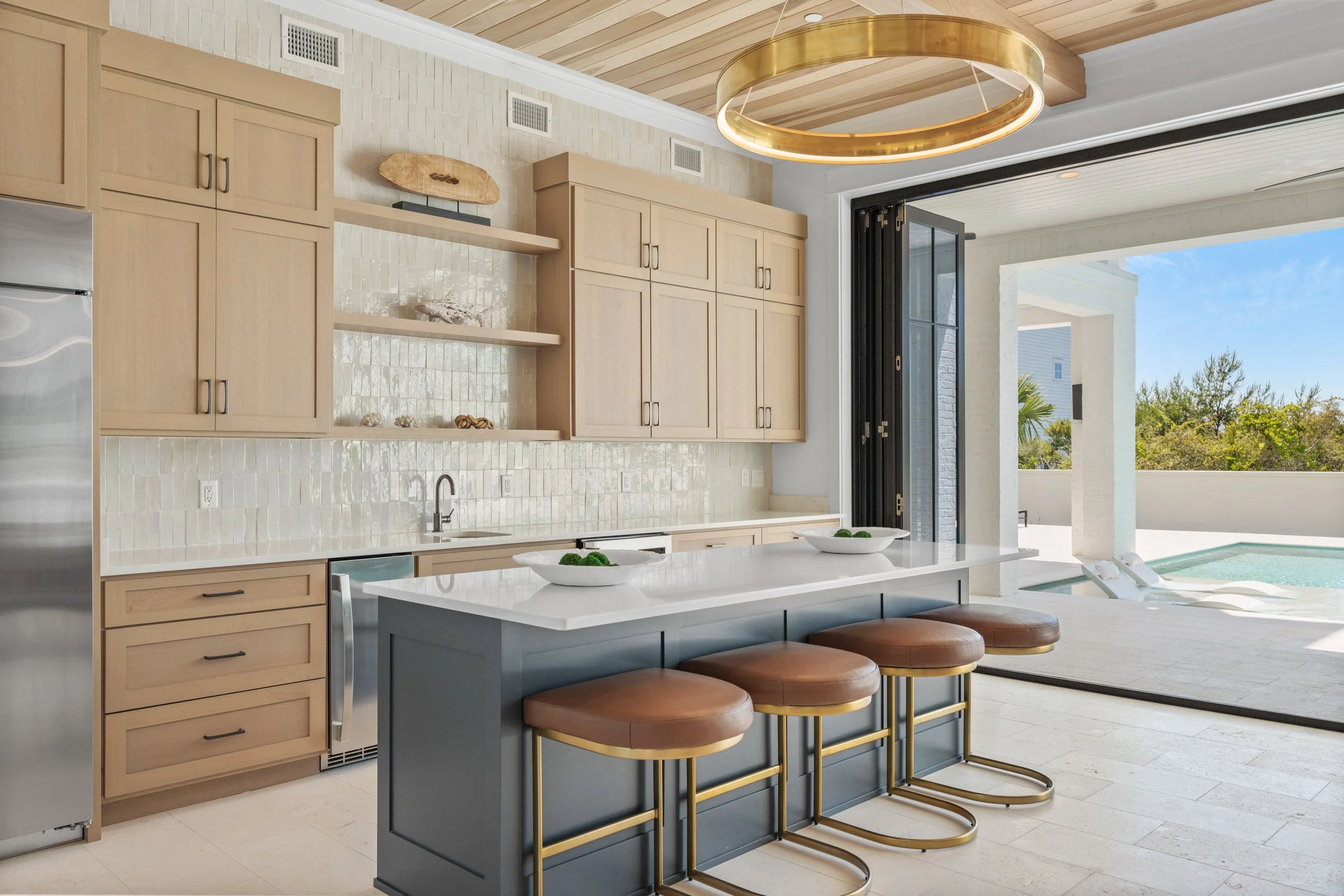 Modern kitchen with beige cabinets, white countertops, and bar stools, opening to a patio and pool area under a blue sky - Real estate photography in 30A, Destin, and Panama City Beach by Matt Jozwiak